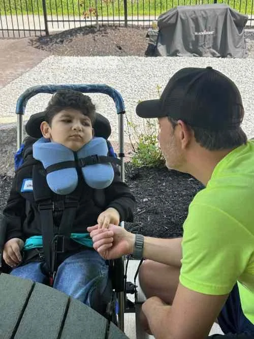 A young boy with a disability sitting in a wheelchair outdoors, holding hands with an adult man wearing a black cap and bright yellow shirt, engaging in conversation, with a backyard setting in the background.