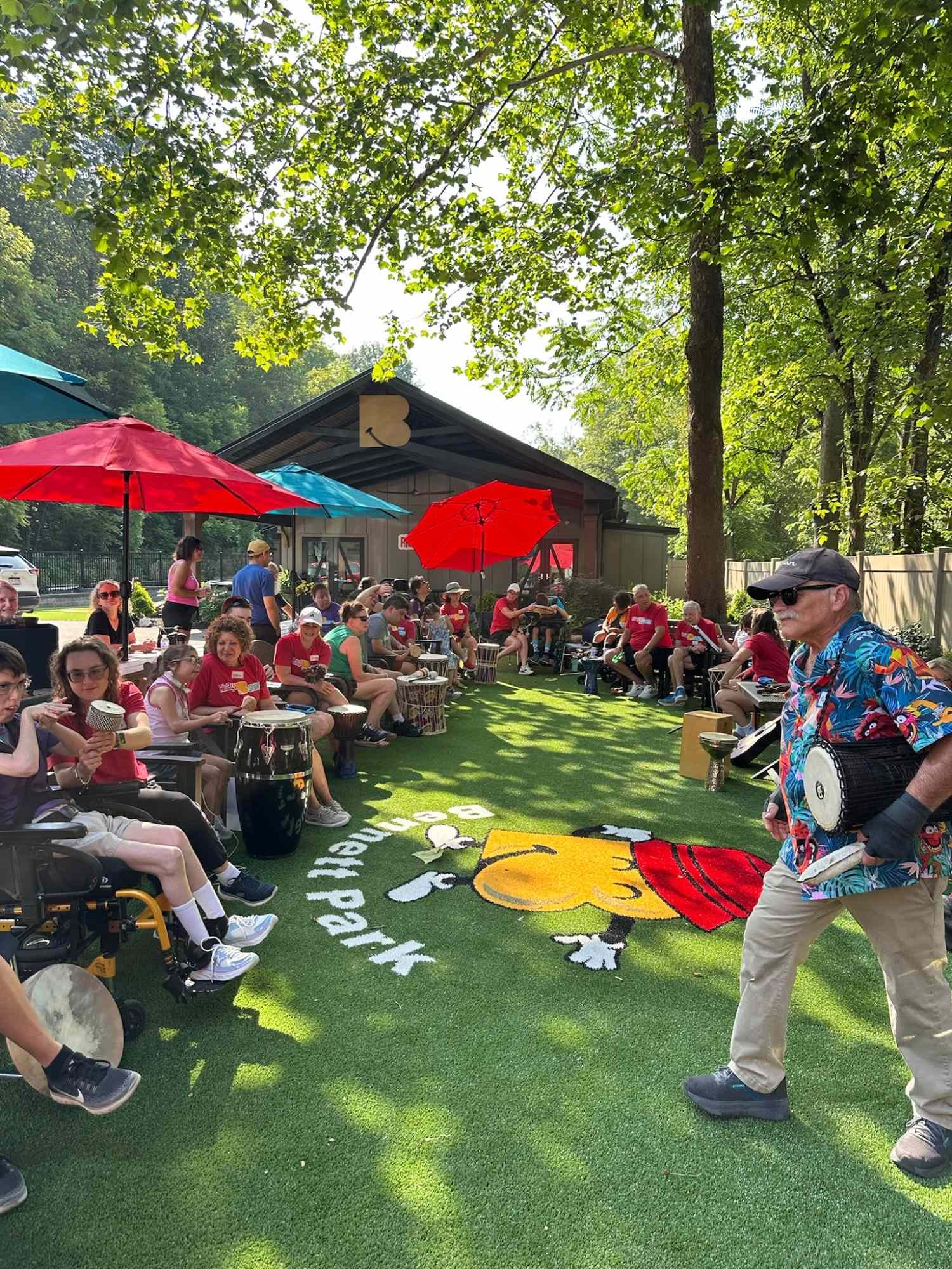 A group of people, including children and elderly, gathered outdoors on a sunny day, sitting around on chairs and drums under red, blue, and teal umbrellas, with a building and trees in the background, and a colorful cartoon character rug on the gras