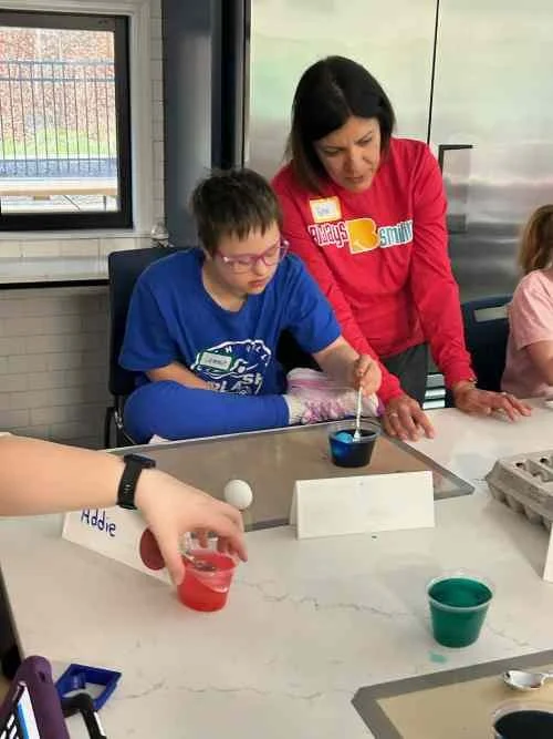 A woman instructing a boy with glasses in a blue shirt during a science activity at a table, with cups, an egg, and a hand holding a small container nearby.