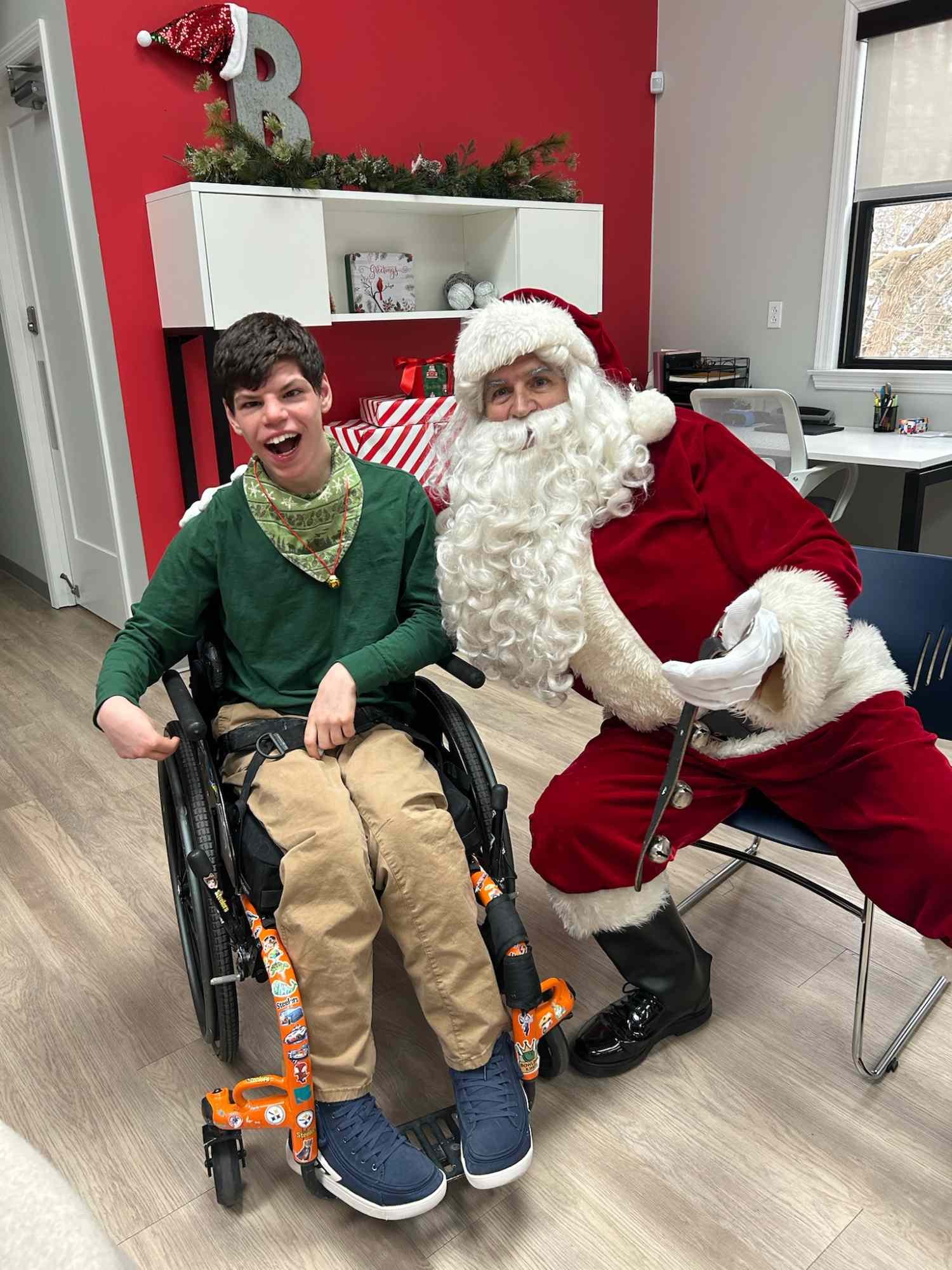 A boy in a wheelchair smiling with a Santa Claus in a red suit and white beard. The room is decorated for Christmas with a red wall, a white shelf with Christmas cards and ornaments, and a Christmas tree with a Santa hat on top.