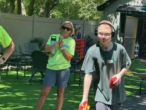A woman and a man in an outdoor backyard setting, with the woman wearing sunglasses and a bright green shirt, and the man wearing glasses and a gray shirt, holding what appears to be gardening tools.