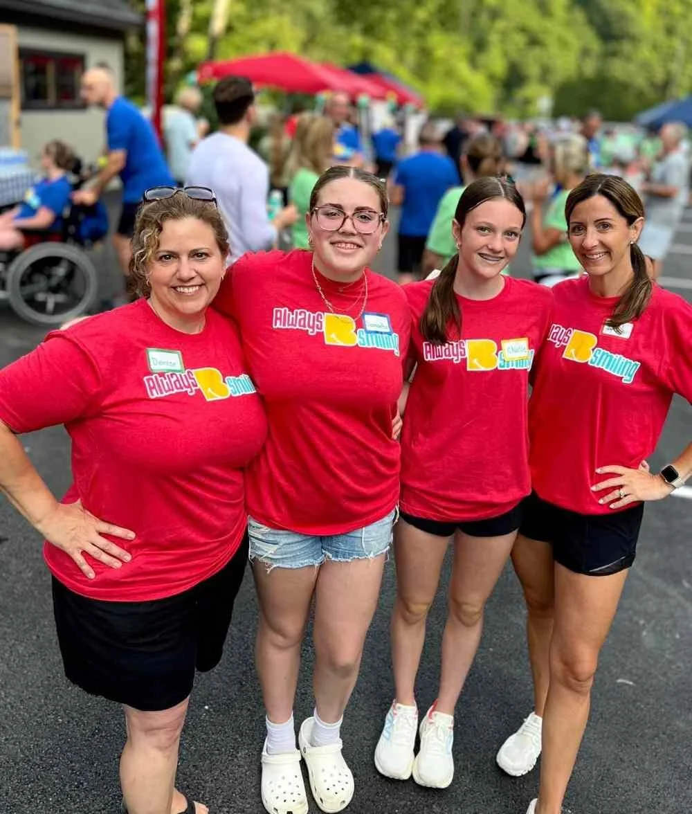Four women wearing matching red T-shirts with the words 'Always Sending' are standing outdoors at a community event, smiling at the camera. They are surrounded by a crowd of people and tents.