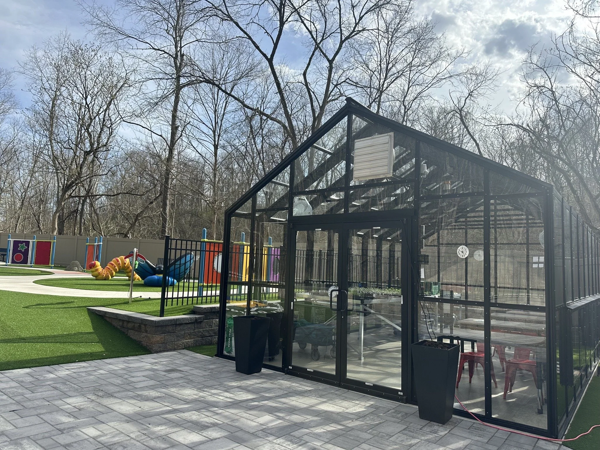 An outdoor playground area with a colorful play structure and a glass-enclosed area, with trees in the background under partly cloudy skies.