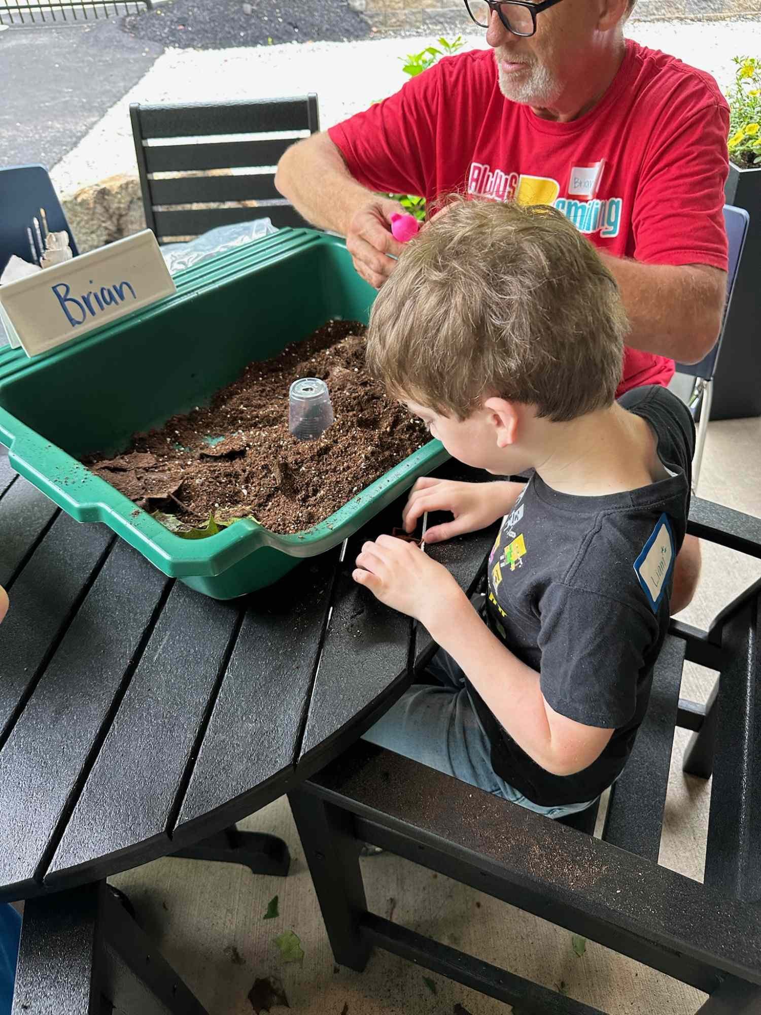A man and a boy planting seeds in a green planter box filled with soil, outdoors on a table.