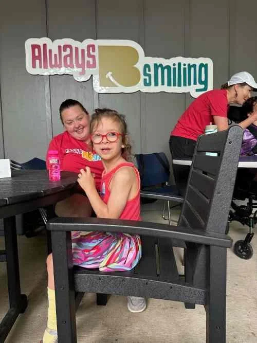 Two young girls sitting at a table with a sign behind them that says, "Always 3 smiling." One girl is wearing glasses and a red shirt, and the other girl is smiling in a red shirt. A woman in a red shirt and white hat is in the background, preparing 