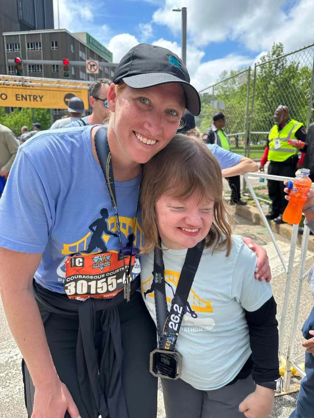 Two smiling people, a woman and a girl, at a marathon event. The woman is wearing a black cap, a blue shirt, and a race bib, while the girl is wearing a light-colored shirt with a medal around her neck. They are outdoors near a fenced area with other runners and event staff in the background.