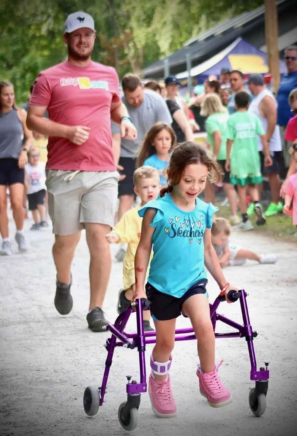 A young girl in a blue Skechers shirt using a purple walker or mobility aid, running with a happy expression at an outdoor event with many children and adults nearby.