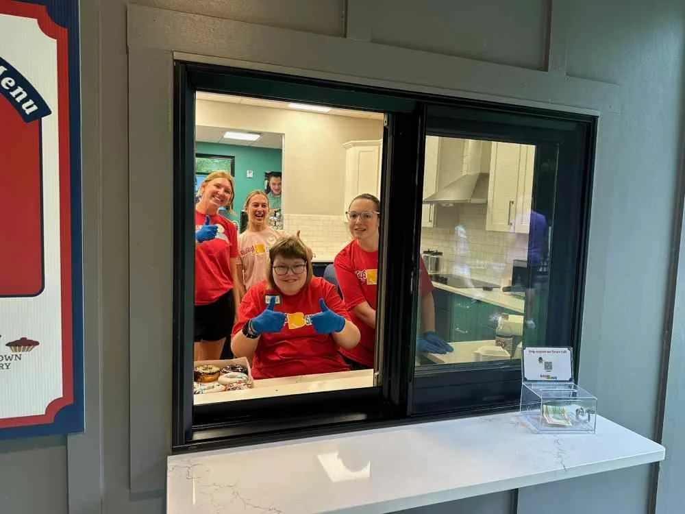 Four smiling people, three women and one young girl, are behind a counter window. They are all wearing red shirts and blue gloves. The girl in front is giving a thumbs-up. The kitchen is visible behind them with white cabinets and a range hood.