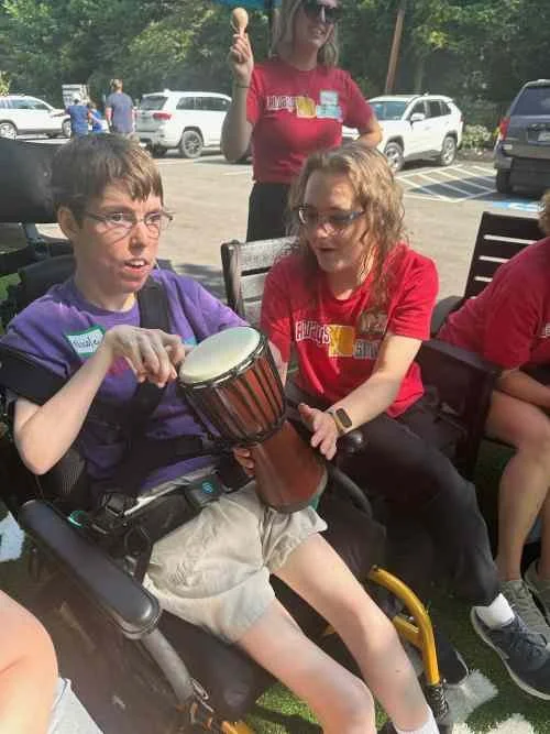 A young person in a wheelchair holding a small drum or hand drum, with two girls nearby smiling and talking, outside in a parking lot with cars and trees in the background.