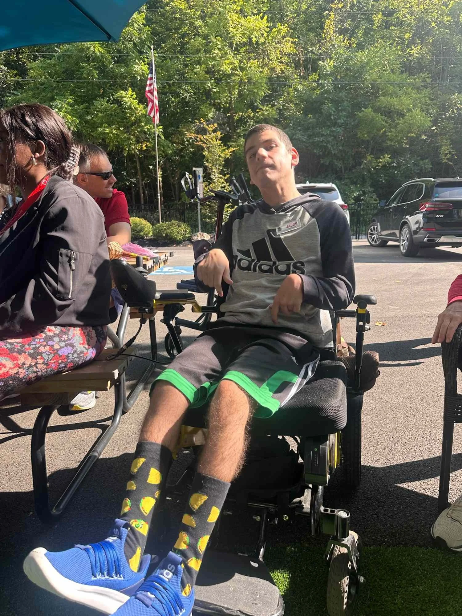 A young man in a wheelchair wearing a gray Adidas hoodie and black shorts with green trim, sitting outdoors on sunny day, surrounded by people, with an American flag and cars in the background.