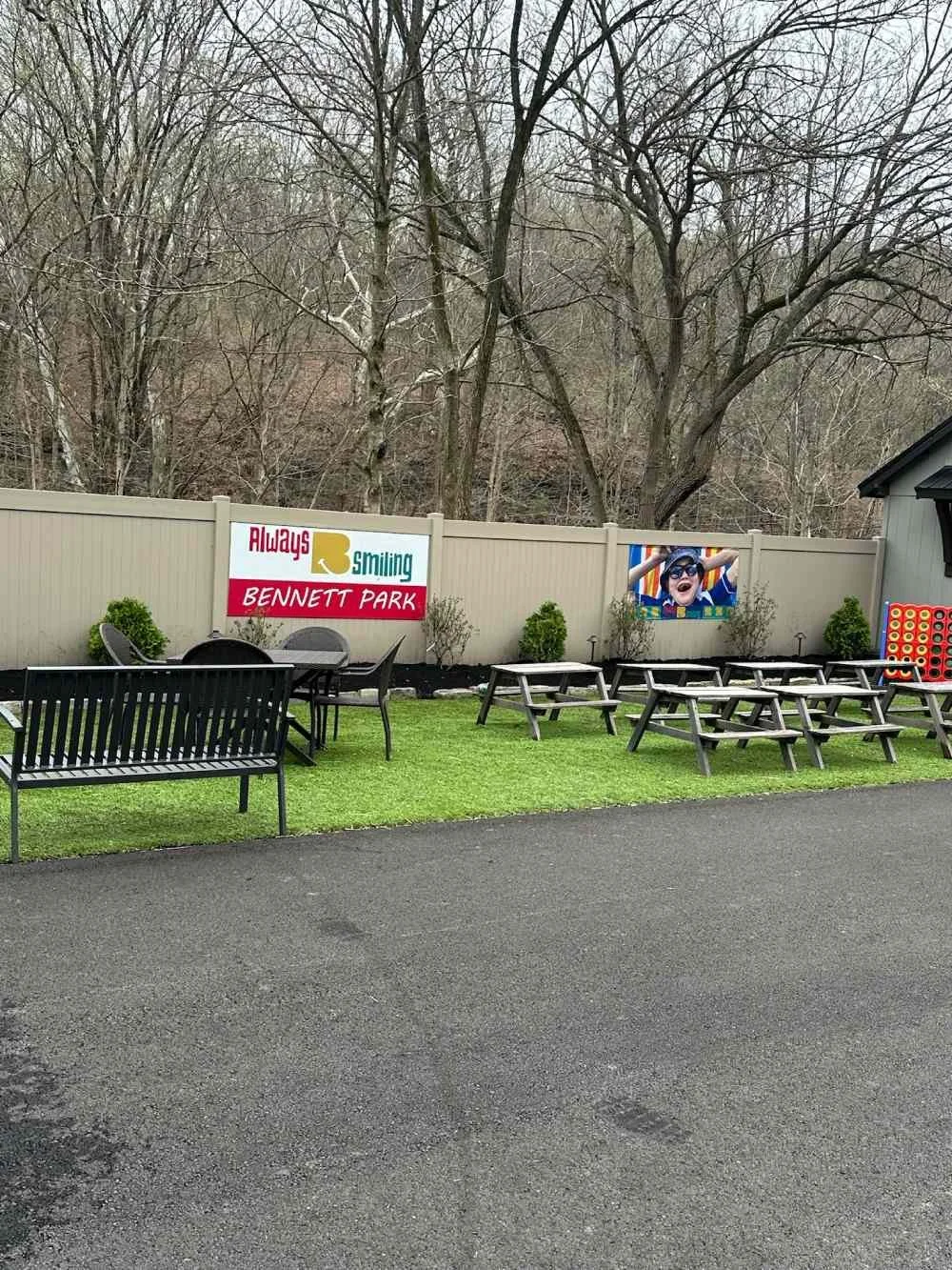 Outdoor seating area with benches and tables in a backyard with a green lawn and a beige fence. There are two signs on the fence, one reading "Always smiling Bennett Park" and a colorful photo of a boy wearing sunglasses and smiling.