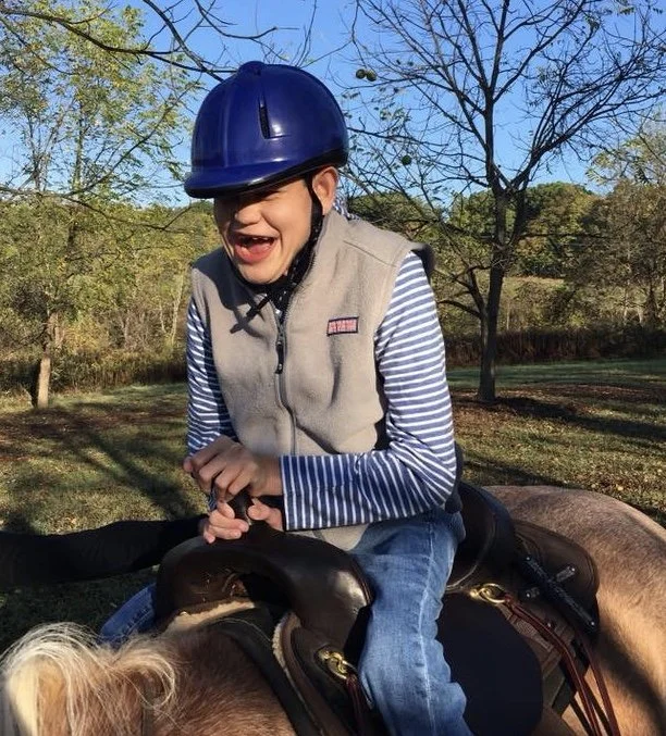 Boy riding a horse outdoors wearing a blue helmet, plaid shirt, and vest, smiling and enjoying the ride on a sunny day.