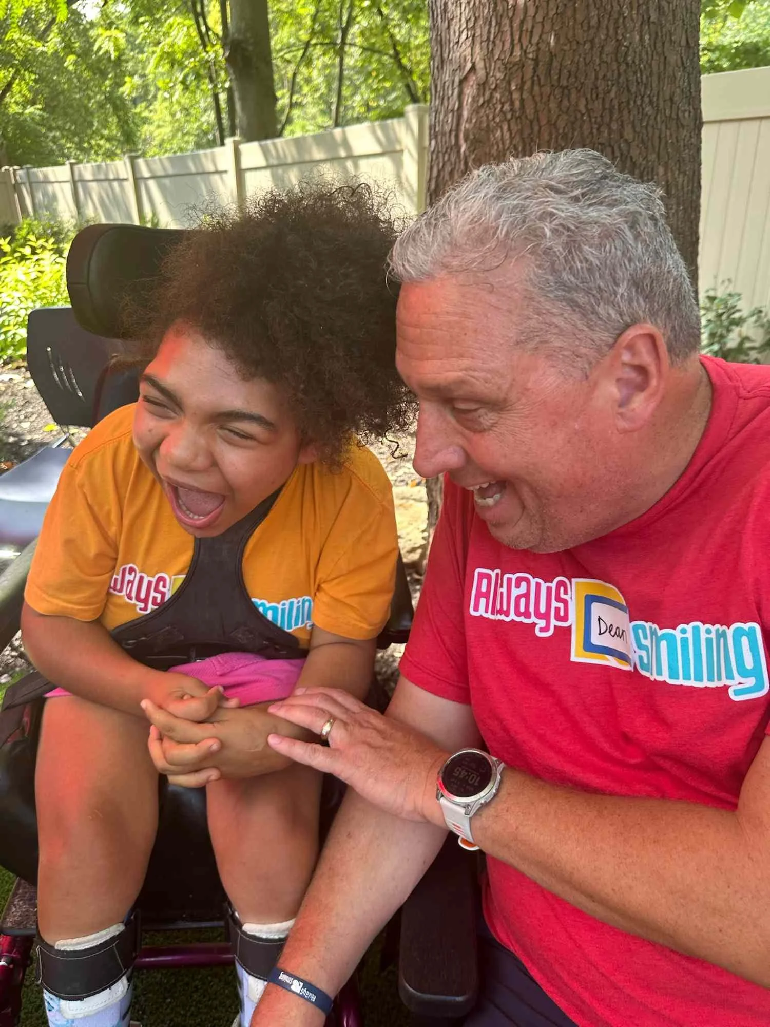 A young girl with curly hair and light brown skin laughing with her eyes closed, sitting outdoors during the daytime. An older man with gray hair, light skin, and a red shirt is smiling and touching her hand, both appearing to share a joyful moment. They are in a green outdoor setting with trees and a white fence in the background.