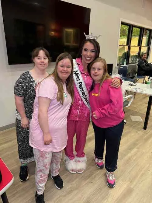 Miss Pennsylvania in a pink outfit wearing a sash, standing with four young girls indoors, smiling for the photo.