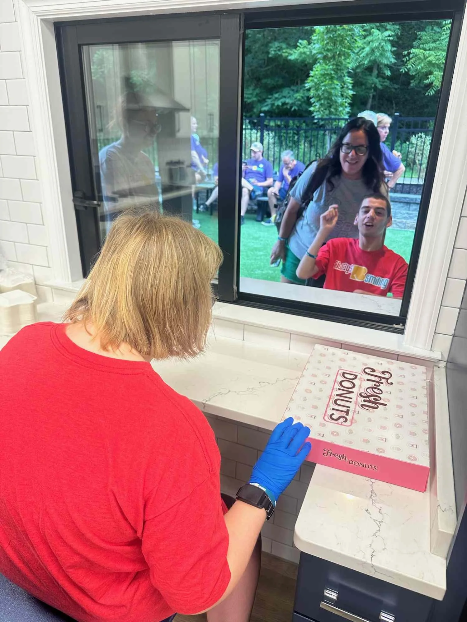 A person in a red shirt and blue gloves standing at a counter with a pink box of Krispy Kreme donuts inside. The person's head with blonde hair is visible from behind. There is a window showing people outside and a woman with dark hair, glasses, and 