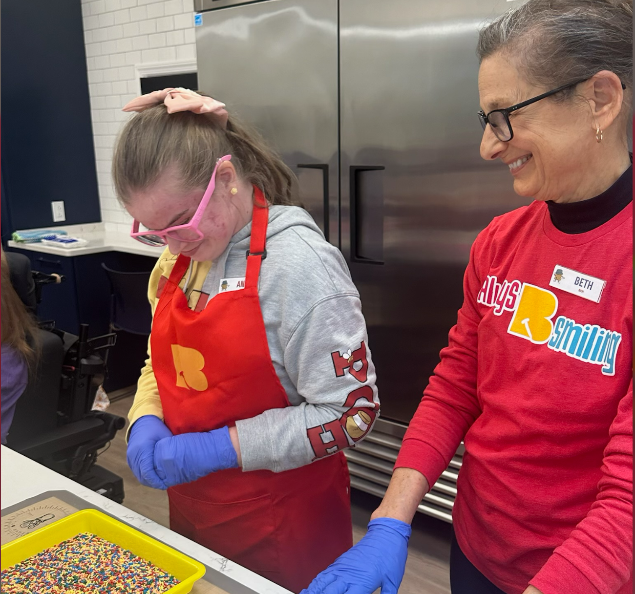 Two women and a girl wearing red shirts and aprons, preparing sprinkles on a tray in a kitchen.