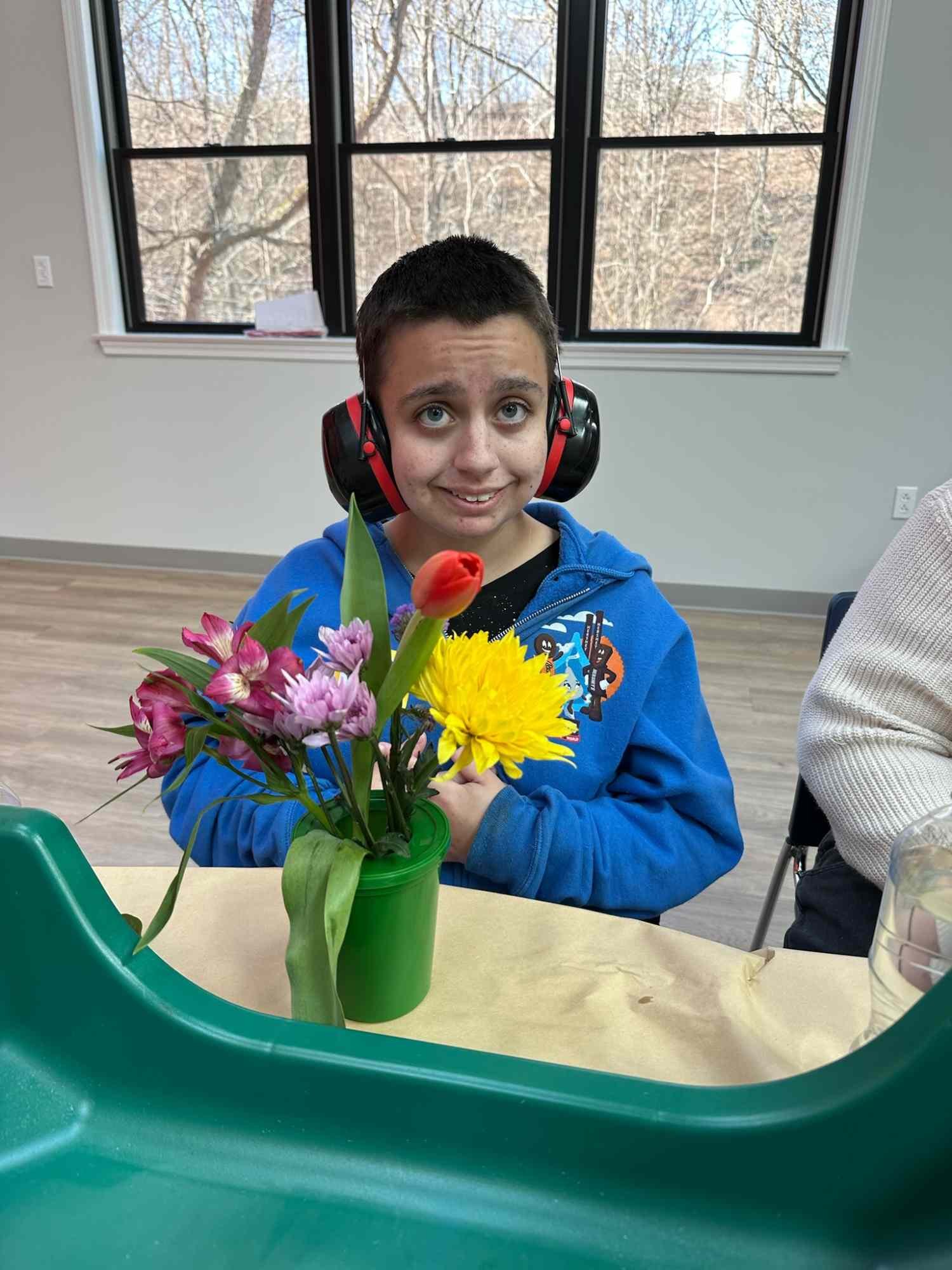 A young boy with short dark hair, blue eyes, and a blue hoodie, wearing large black and red noise-canceling headphones, is sitting at a table with a green flower pot of colorful flowers, including yellow, pink, purple, and red blooms, in front of him