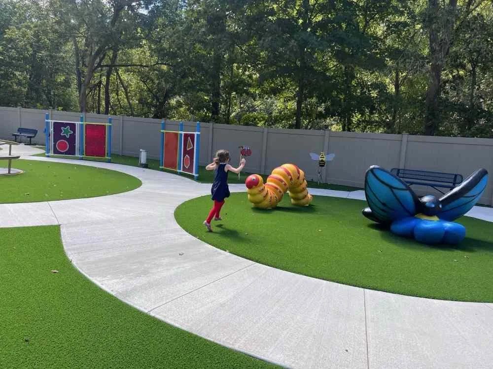 A young girl playing in a colorful outdoor playground with worm, bee, and butterfly sculptures, and artificial grass, enclosed by a gray fence, with tall trees in the background.