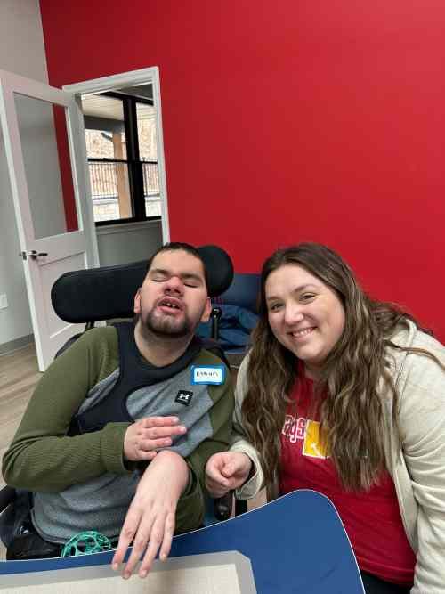 A young man with a disability in a wheelchair is sitting next to a woman with long brown hair, both smiling. The background features a red wall and a doorway leading to a room with a window.