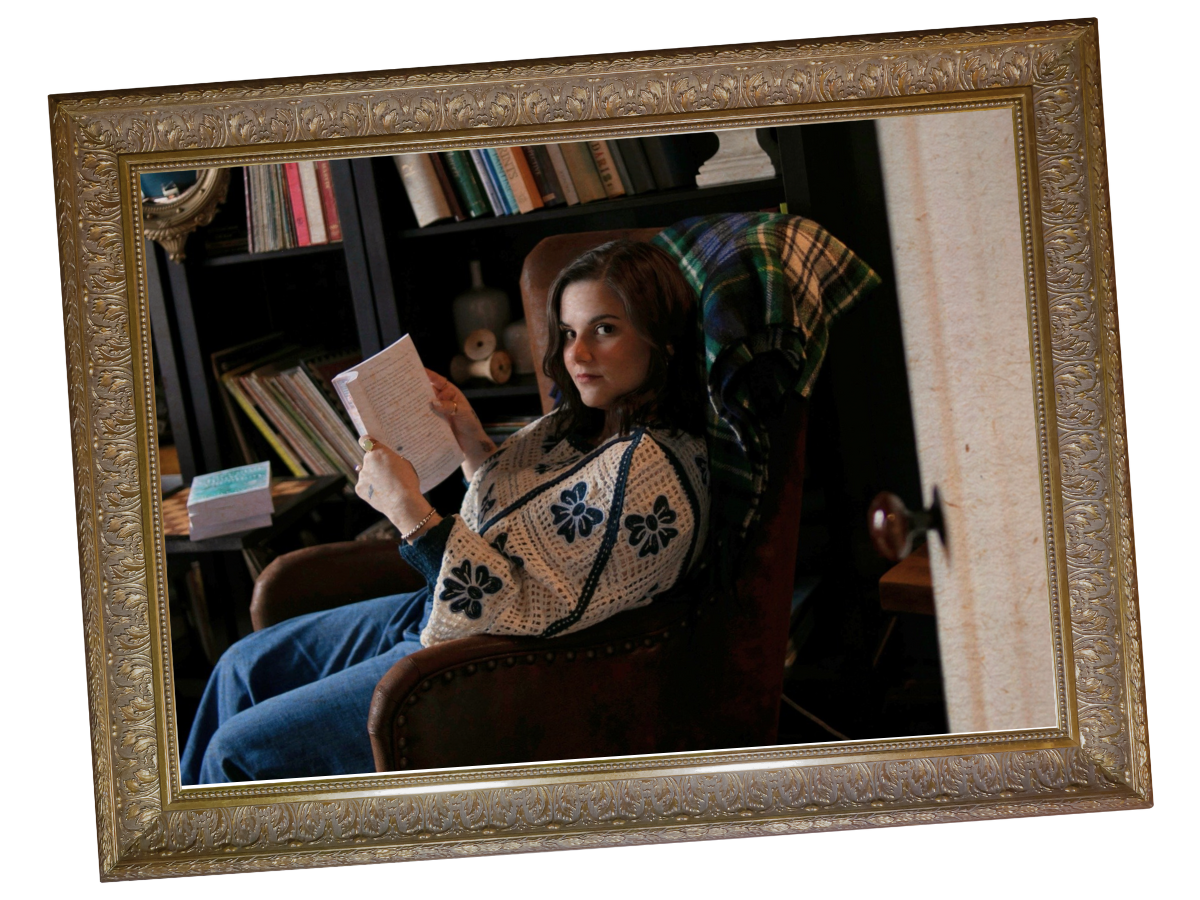 A woman, the author, with dark hair sitting in a brown armchair, holding an open book, with bookshelves and decorative items behind her, and a plaid blanket draped over the chair.
