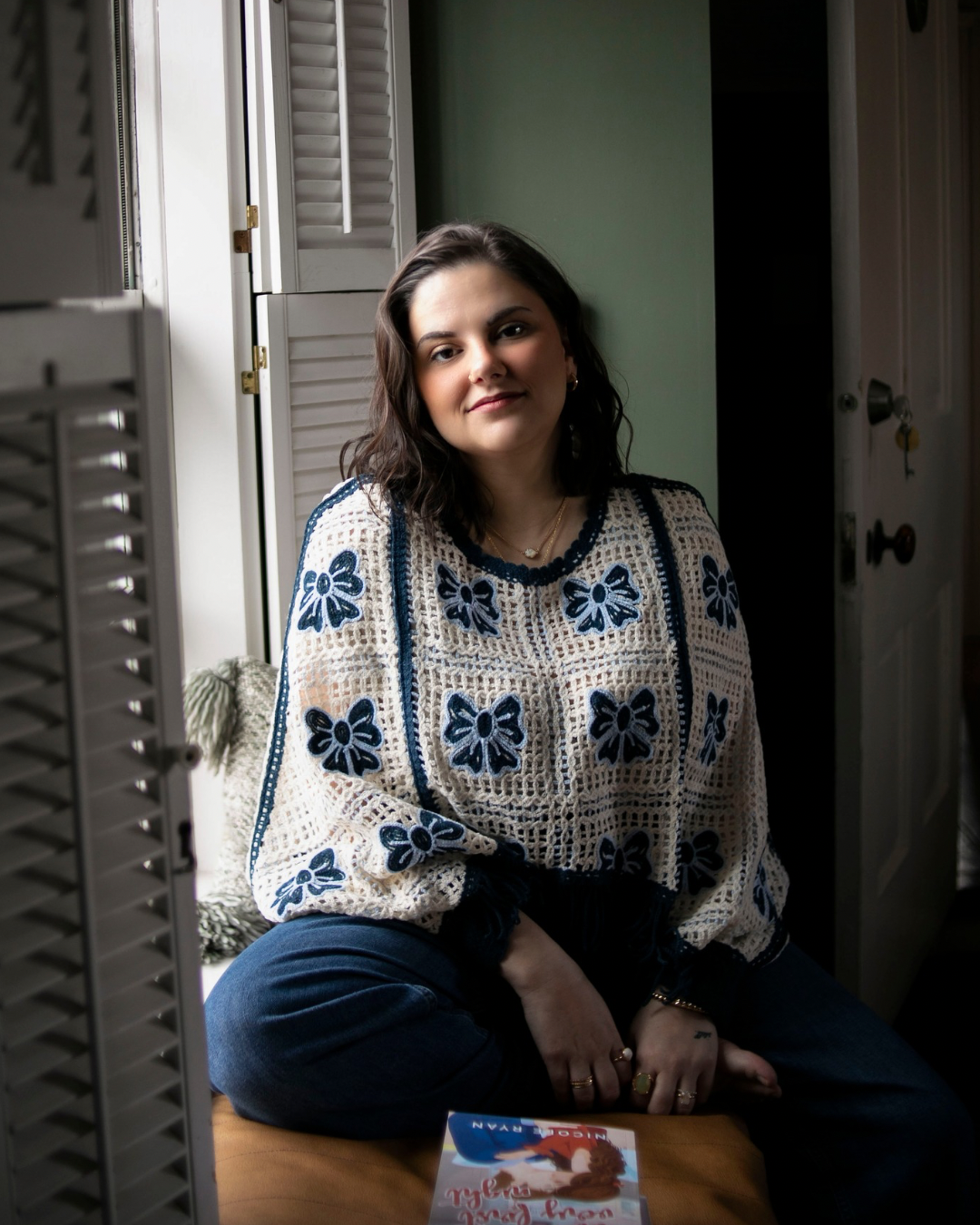 A young woman with dark wavy hair sitting on a wooden surface near a window with open white shutters, wearing a cream-colored crochet sweater with blue floral embroidery, and dark jeans.