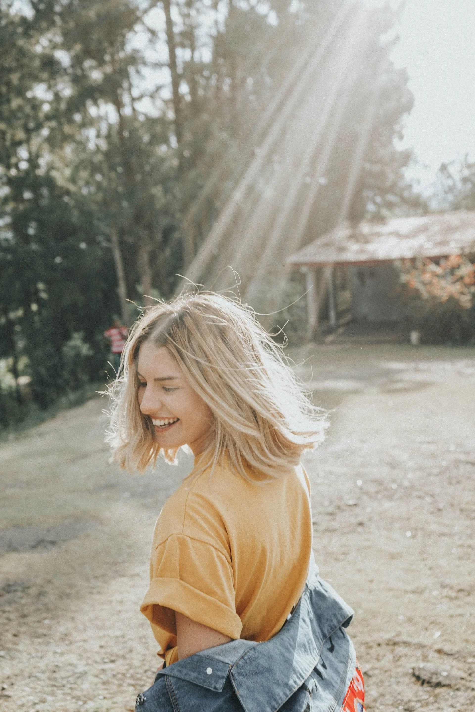 A young woman with blonde hair smiling as she looks downward, sunlight shining down on her from the upper right, in an outdoor park setting with trees and a wooden shelter in the background.