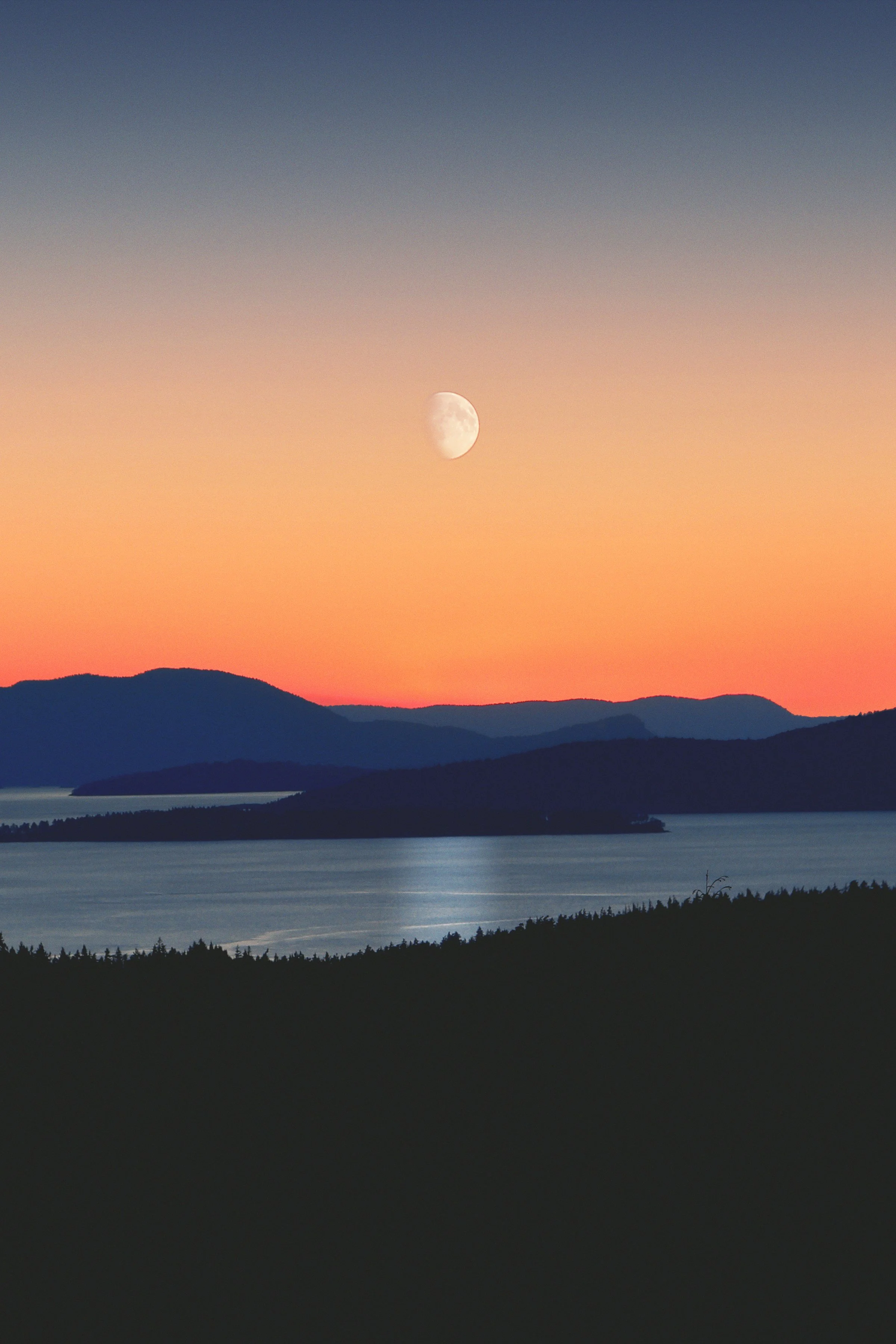 A landscape photo capturing a lake with mountains in the background during sunset, a visible moon in the sky, and a gradient of colors from orange to blue.