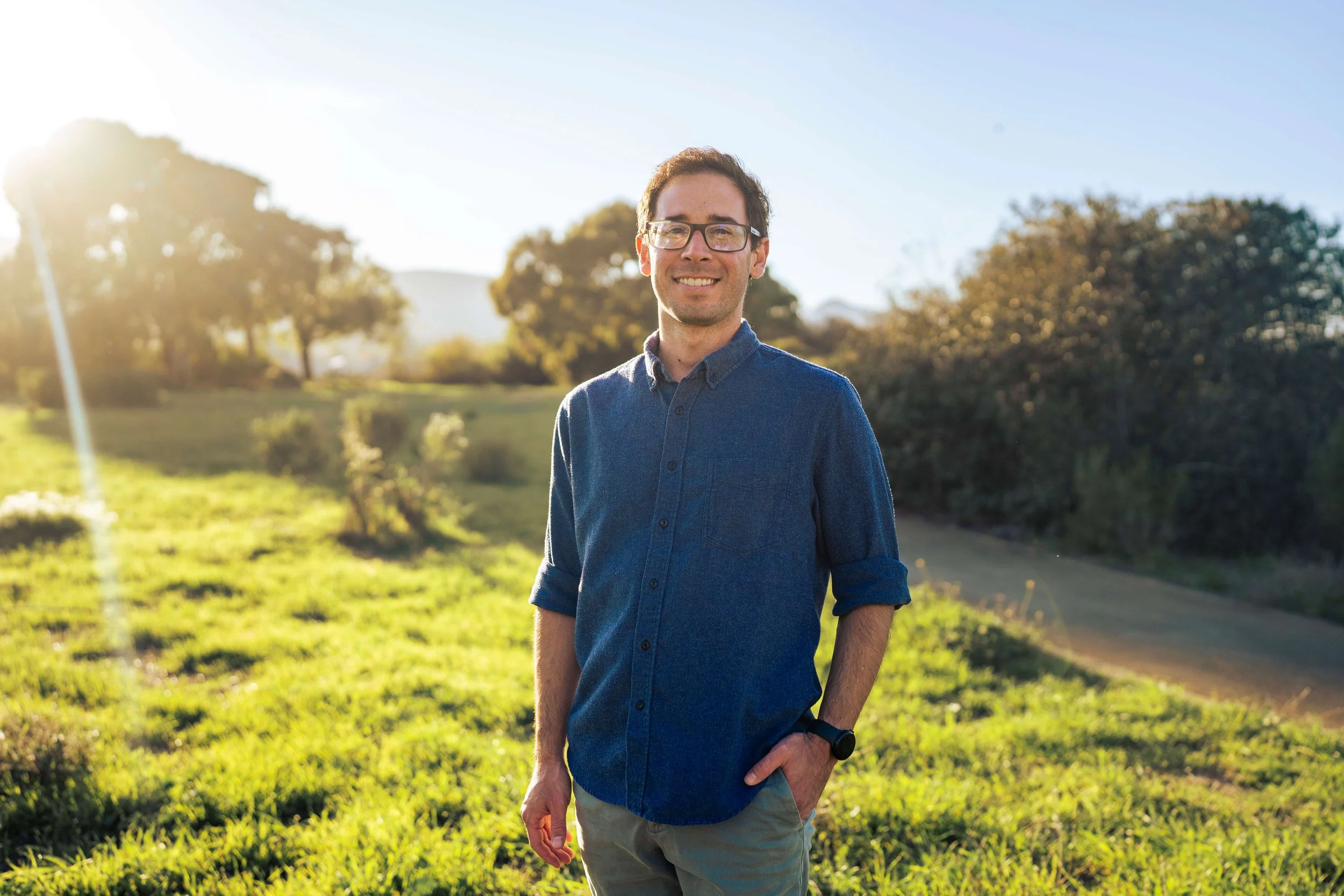 A smiling man with glasses, wearing a blue shirt and beige pants, standing outdoors in a sunny park or field with trees and grass in the background.
