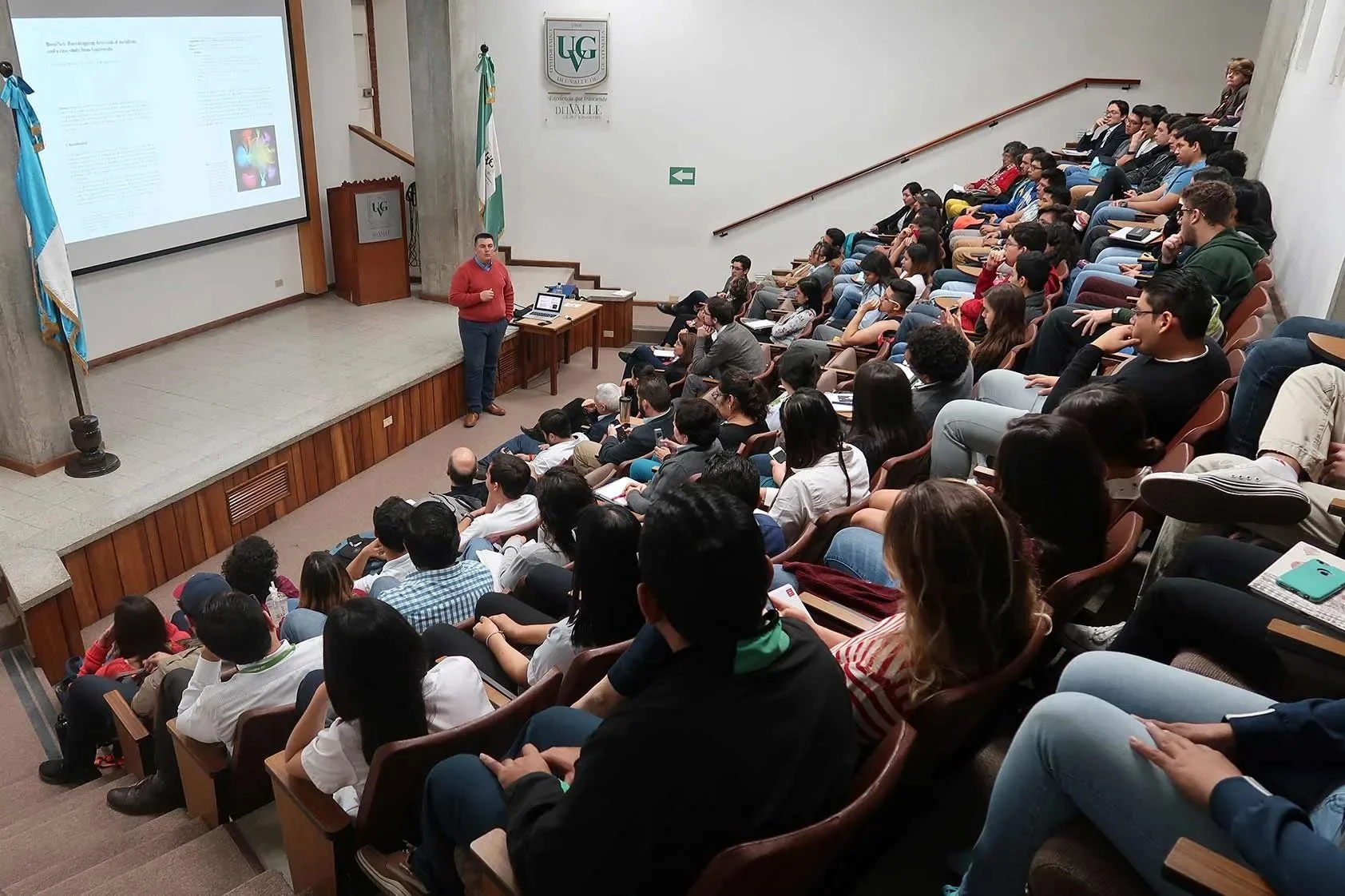 A lecture hall filled with students listening to a speaker at the front of the room, with a presentation projected on a large screen.