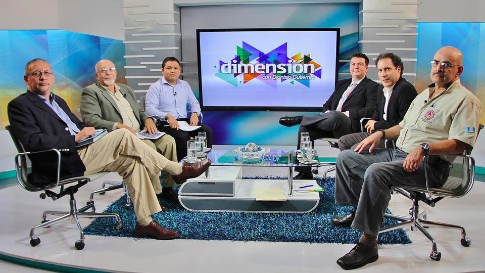 Six men sitting around a glass table in a television studio with a large screen behind them displaying the word "dimensión". The studio has modern decor and a colorful background.