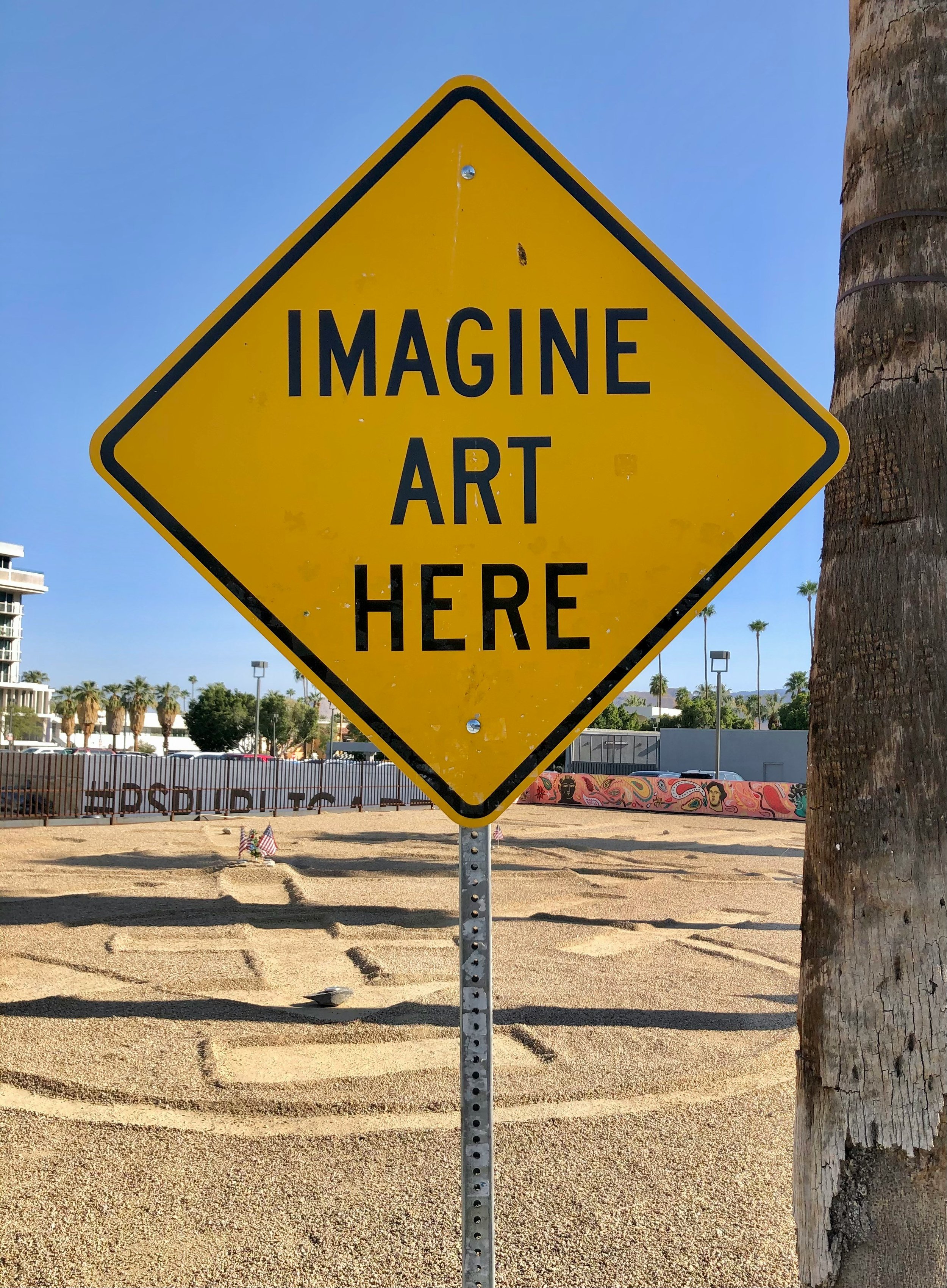 Yellow diamond-shaped sign with black border and black text that reads 'IMAGINE ART HERE' mounted on a metal pole next to a tree, with a background of a park or public space with graffiti, trees, and a clear blue sky.