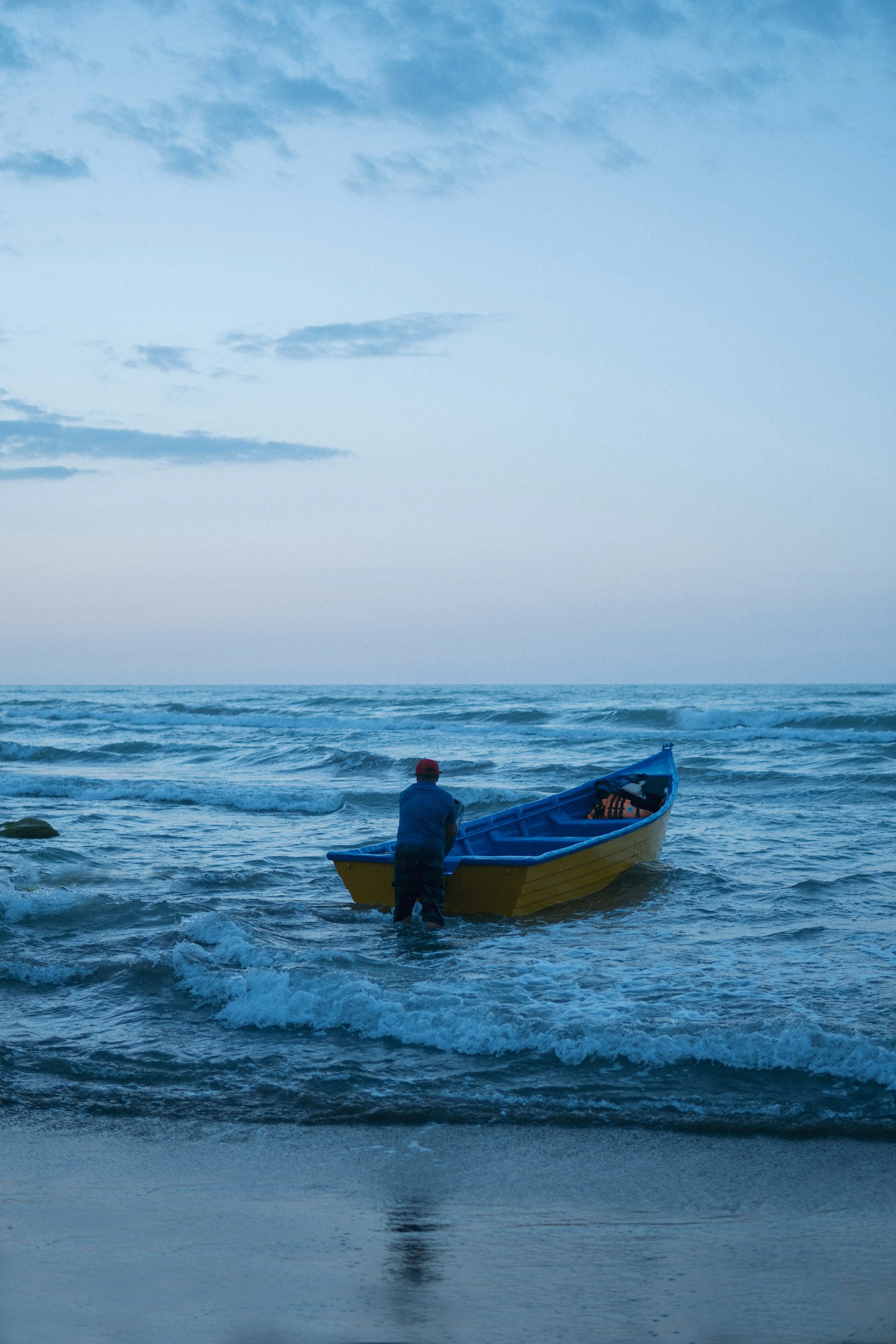 A person with a red cap standing in the water next to a yellow and blue boat at the shoreline, with the ocean and cloudy sky in the background.