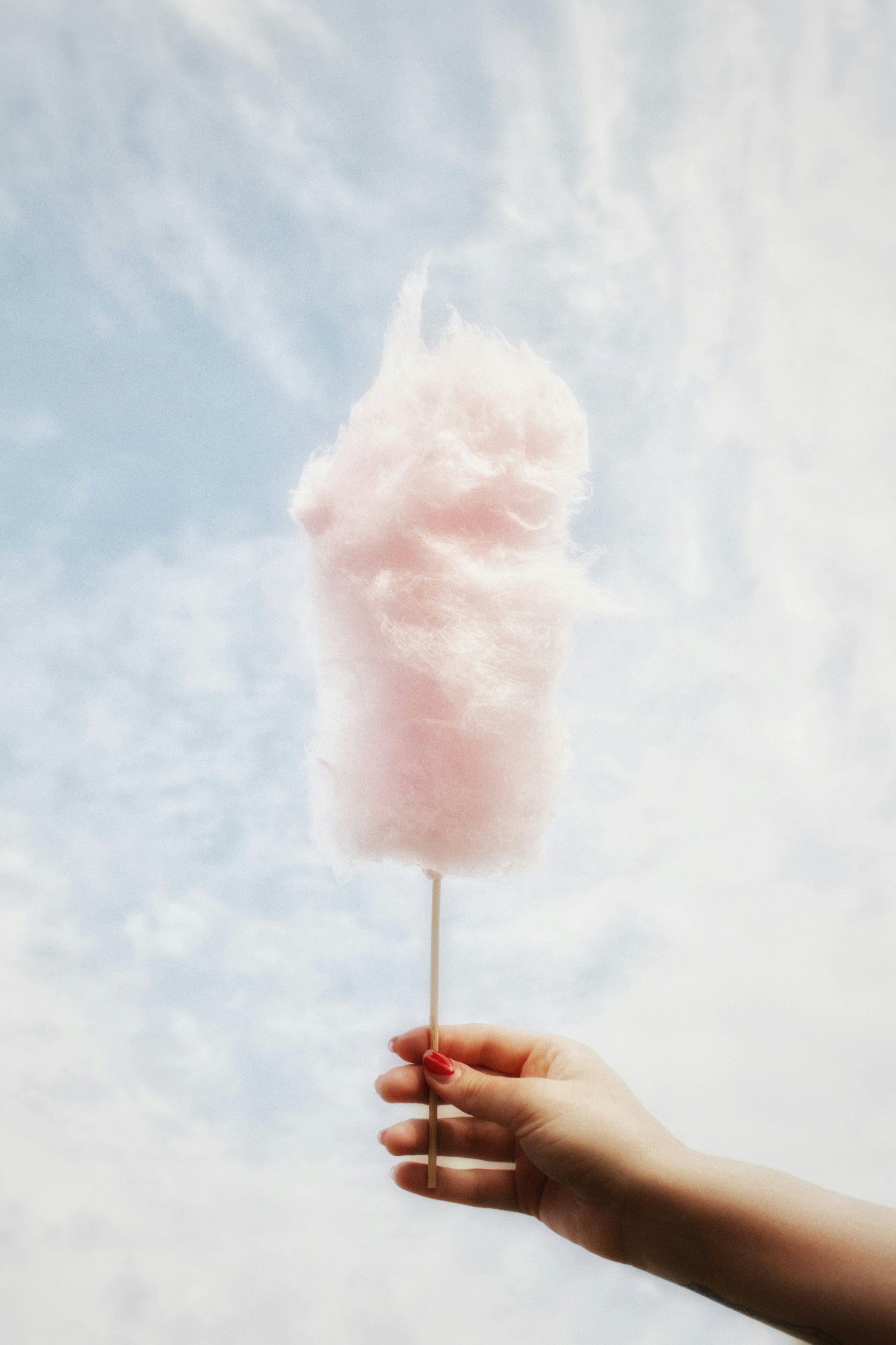 A hand holding a pink cotton candy on a stick against a cloudy sky background.