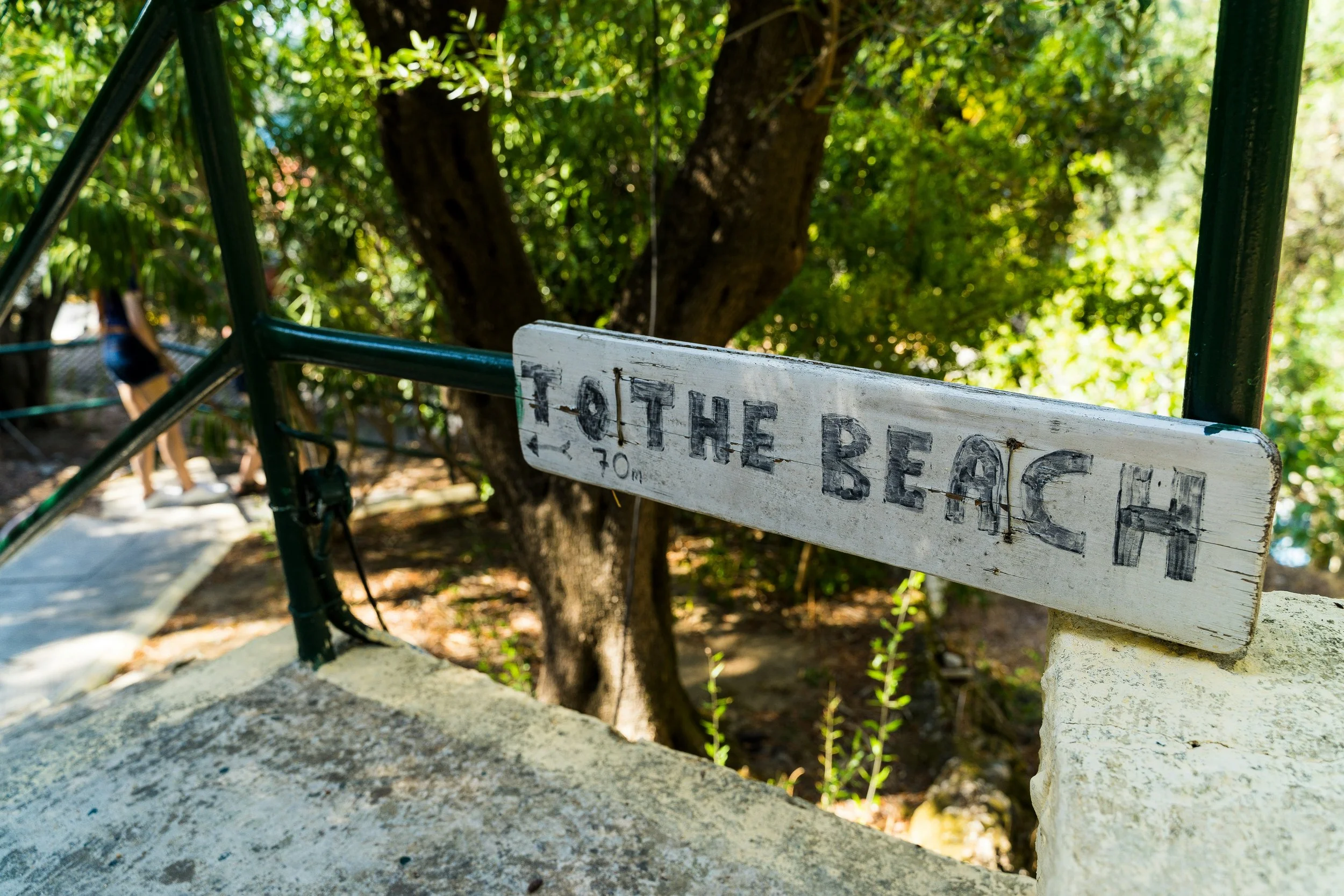 Close-up of a weathered white wooden signpost reading 'THE BEACH', attached to a black metal railing by some twine, with a sandy pathway, green trees, and a person in the background.