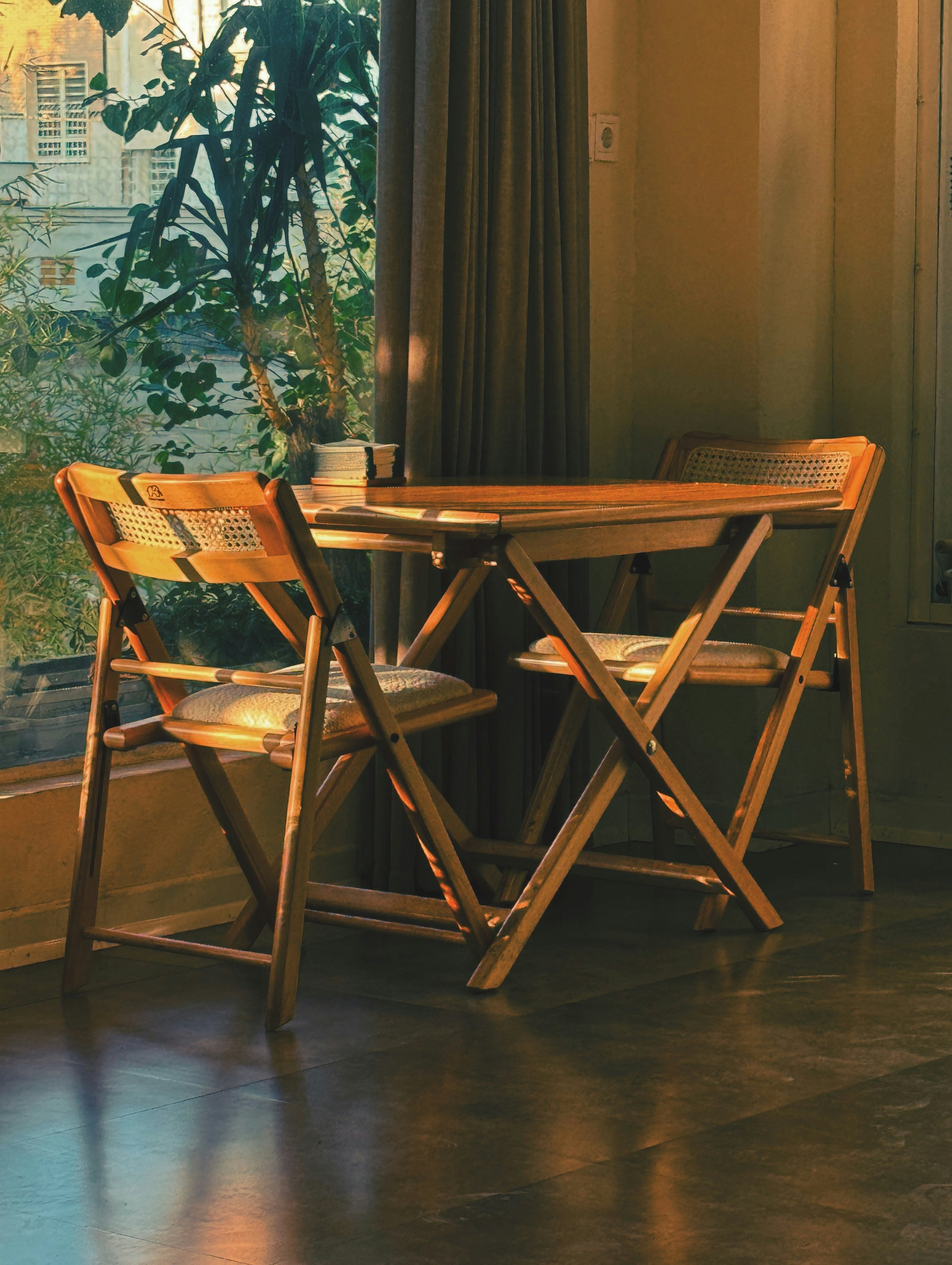 A wooden dining table and chairs near a window with sunlight and a view of green plants outside.