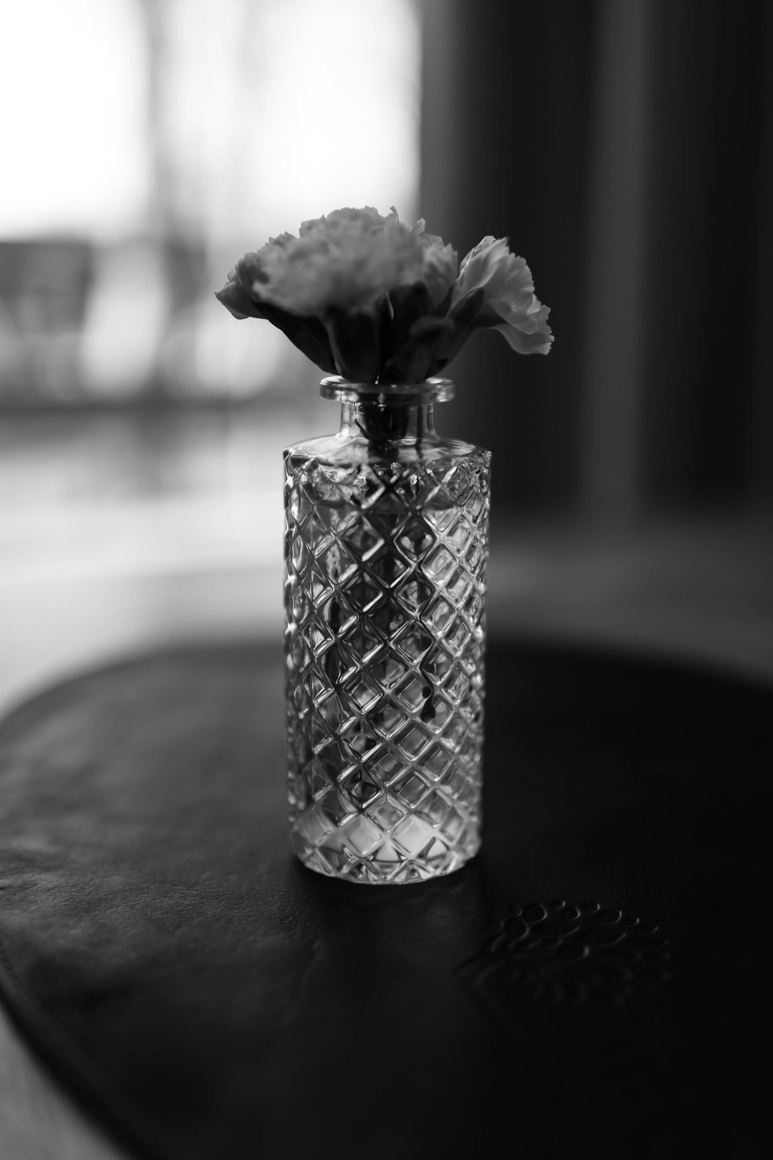 A black and white photo of a small, ornate glass vase with a textured diamond pattern, holding a single flower, placed on a dark surface.