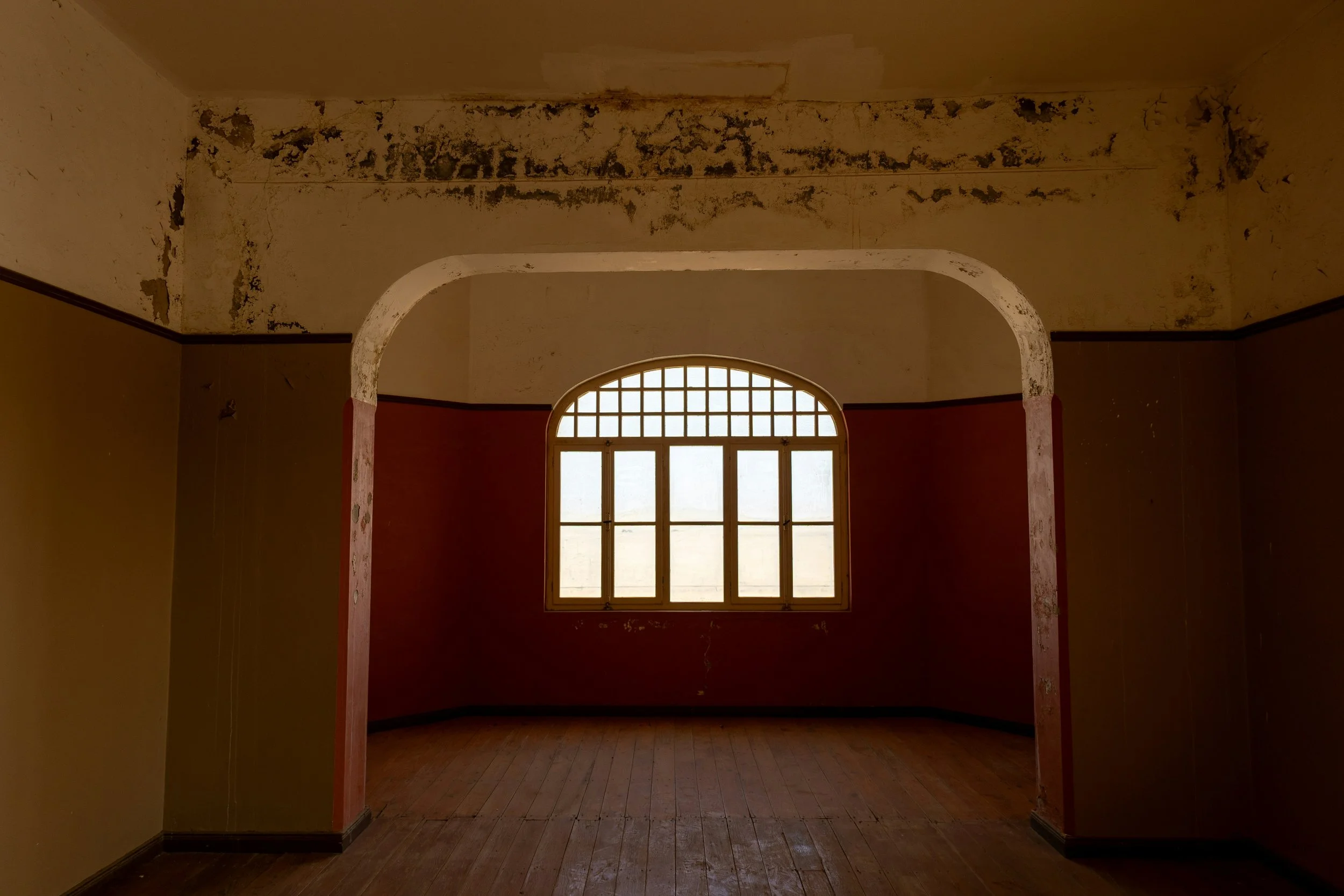 An empty room with peeling paint on the ceiling and walls, a large arched window with frosted glass, and hardwood floors.