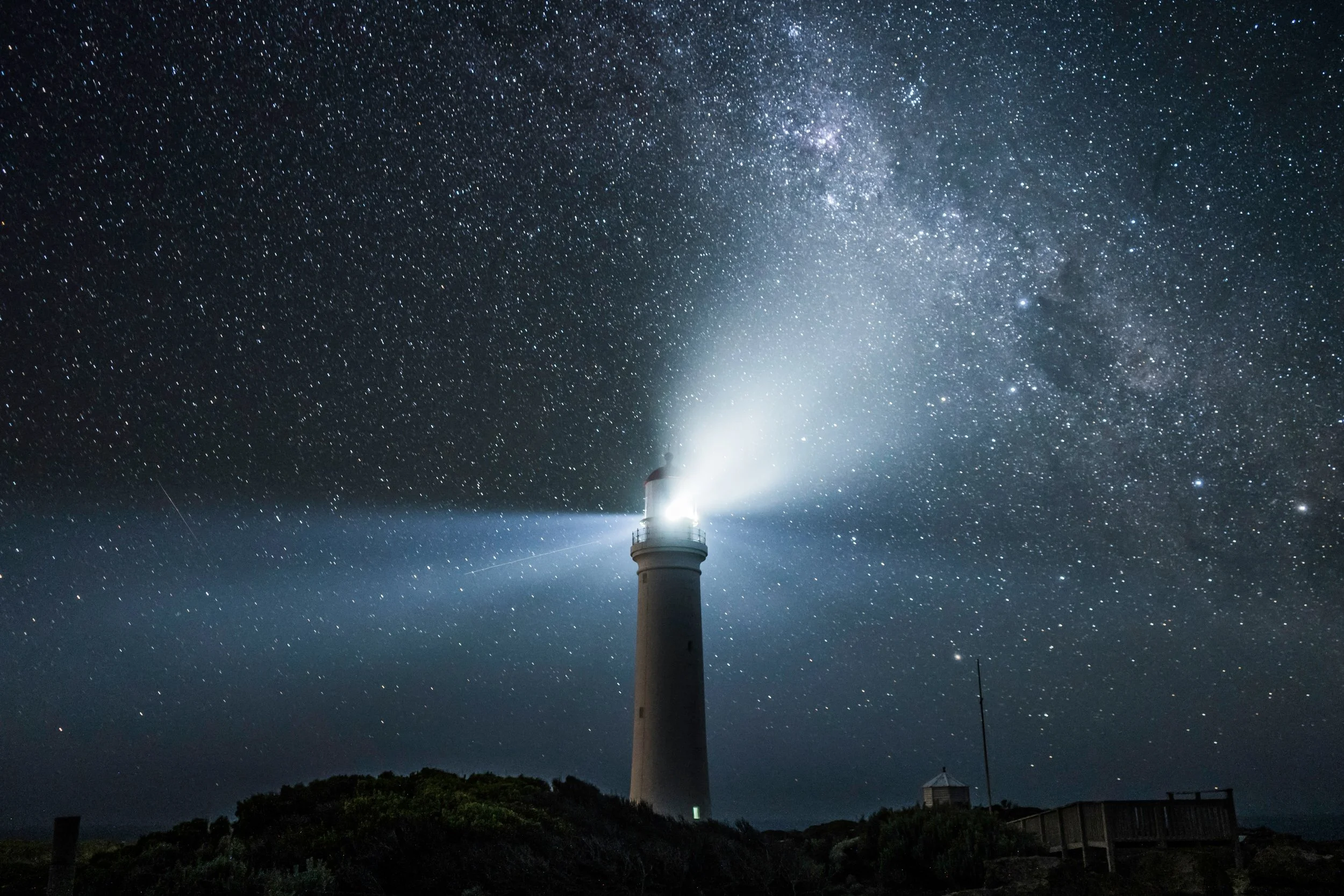 A lighthouse shining bright light into the starry night sky, with the Milky Way visible above.