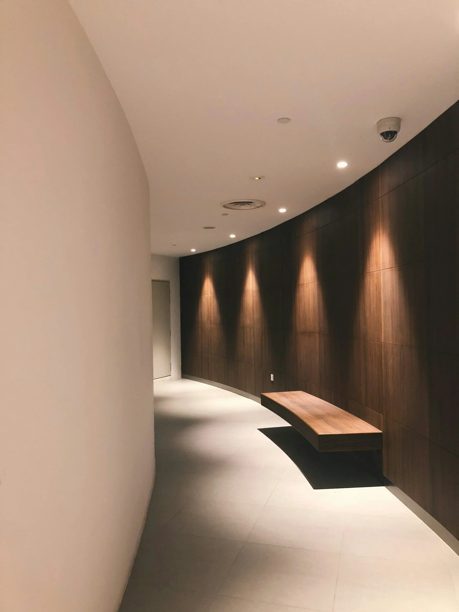 Empty hallway with a curved white wall on the left and a dark wood-paneled wall on the right, with a wooden bench attached to the dark wall. Ceiling has small recessed lights. Drywall.