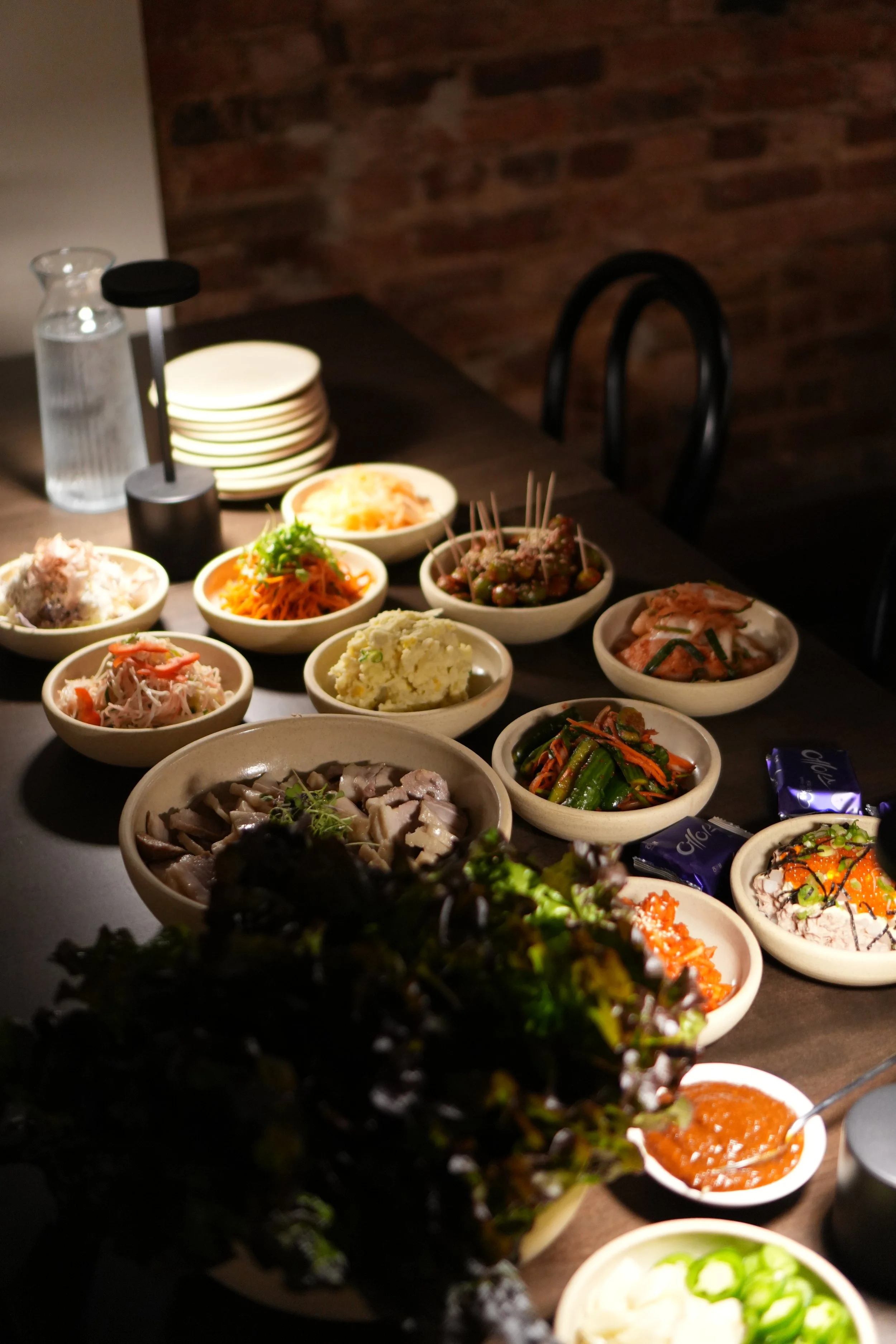 A variety of Korean dishes served in small bowls on a dark table, with a brick wall in the background at Bar Alice Anne.