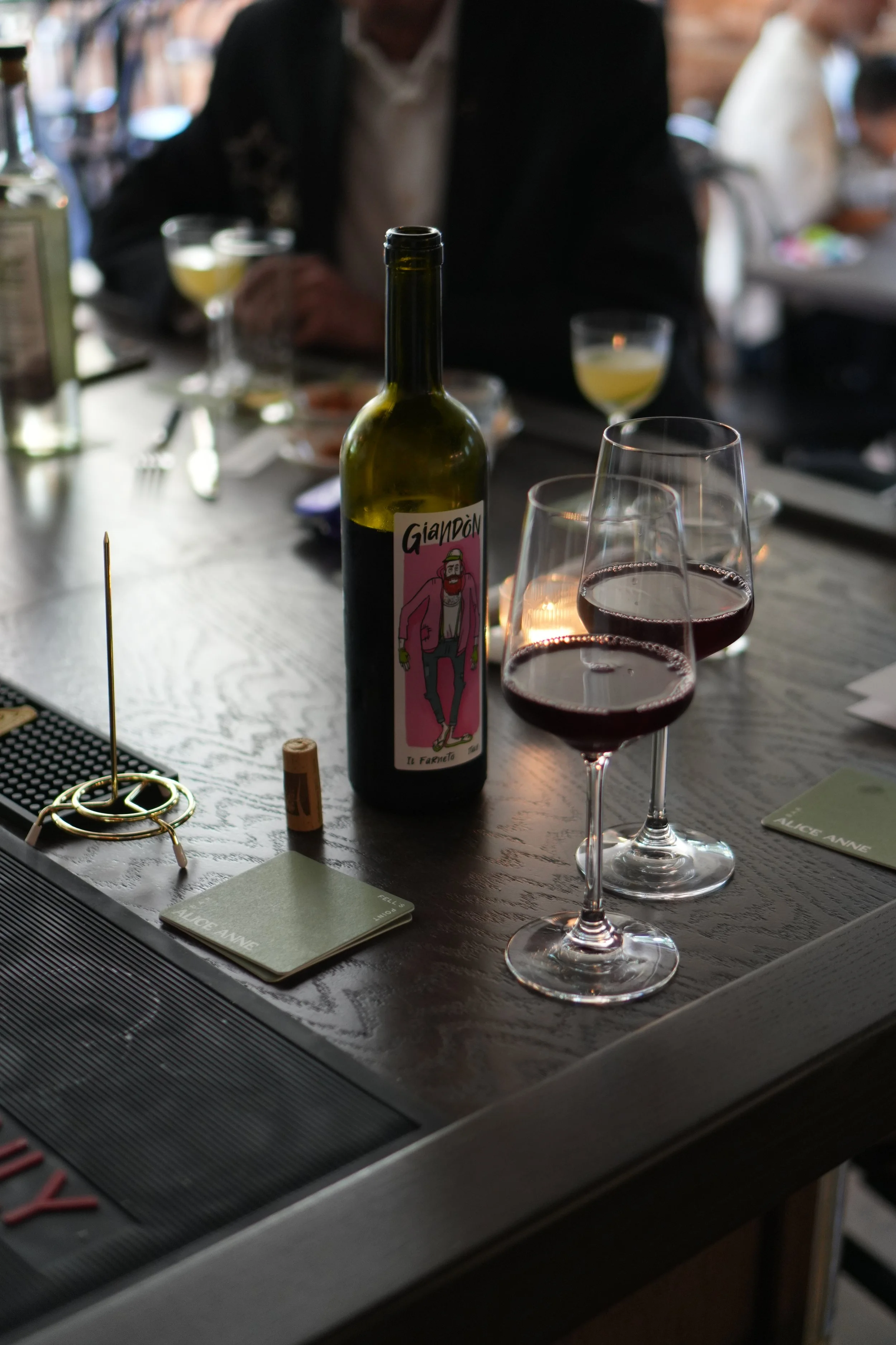 A bottle of wine, three glasses of red and white wine, and a wine cork on a table at Bar Alice Anne. In the background, a man is sitting with two glasses of white wine, and other people are visible.
