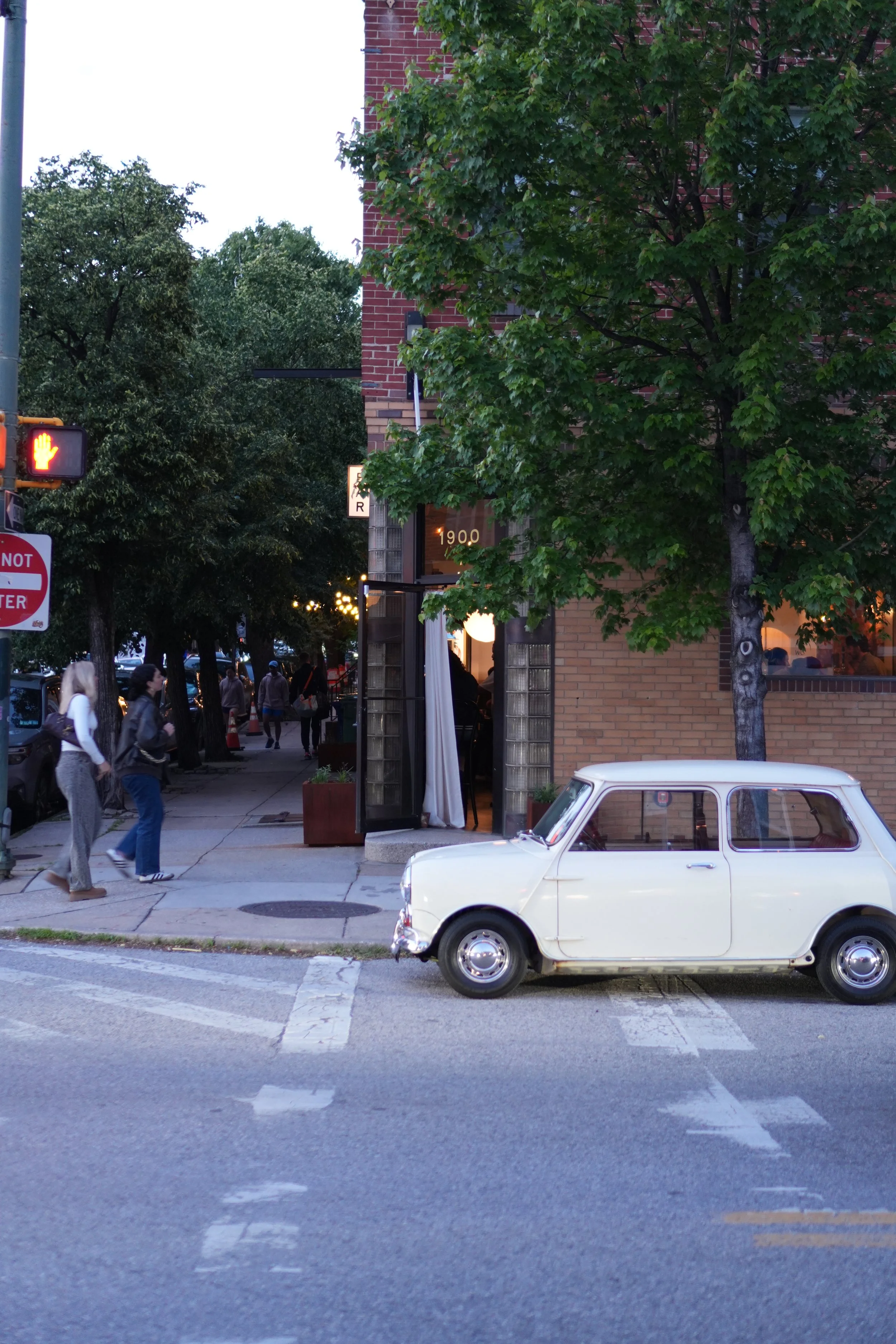 Bar Alice Anne and Street scene with pedestrians walking on the sidewalk, a parked vintage white car, a brick building with green trees in front, and a traffic light showing a red hand signal.