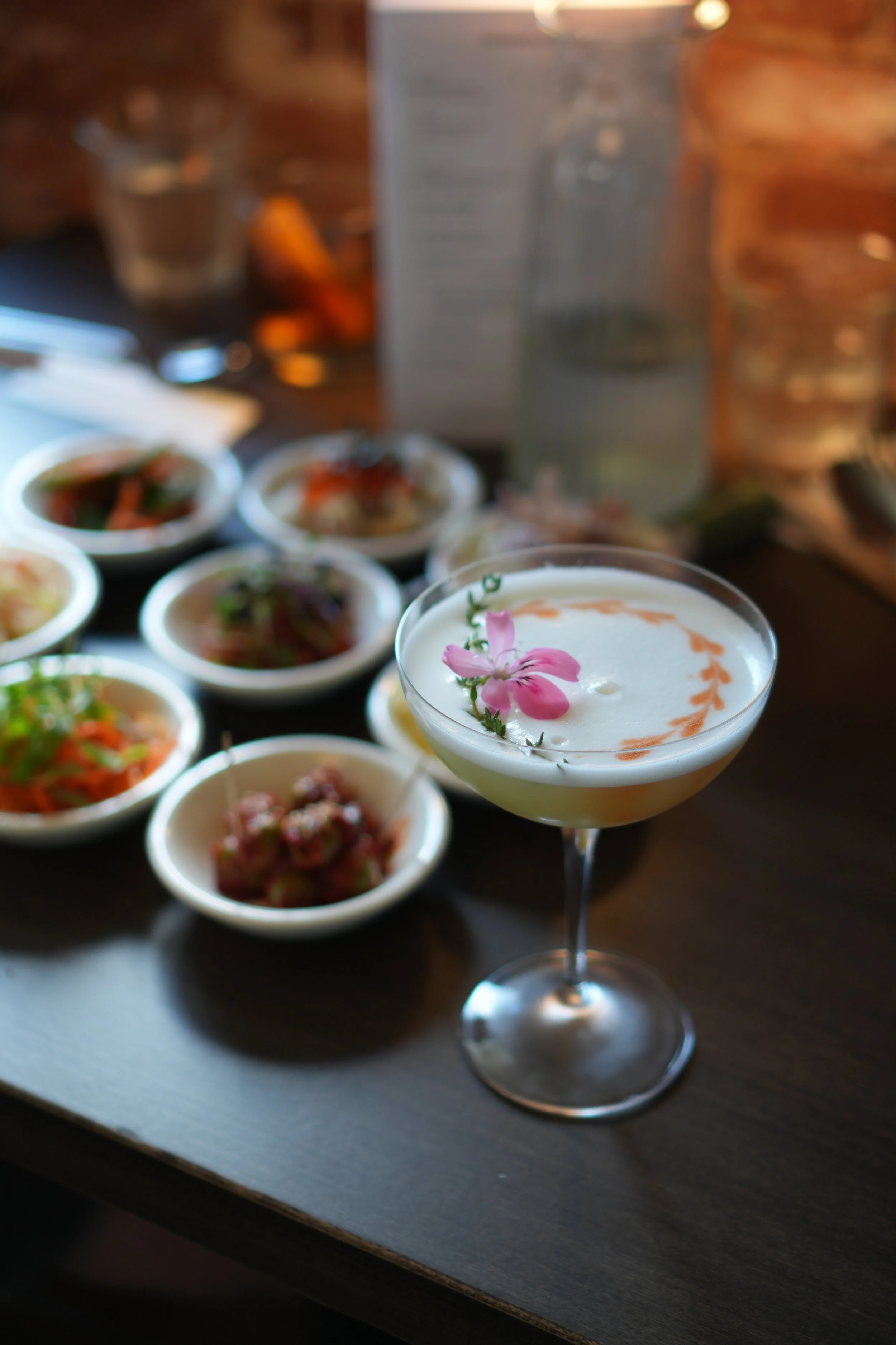 A cocktail glass with a pink flower and greenery garnish in the foreground, with several small bowls of various dishes on a dark table in the background, some of which contain vegetables or meats, and a paper menu and glass of water are also visible.