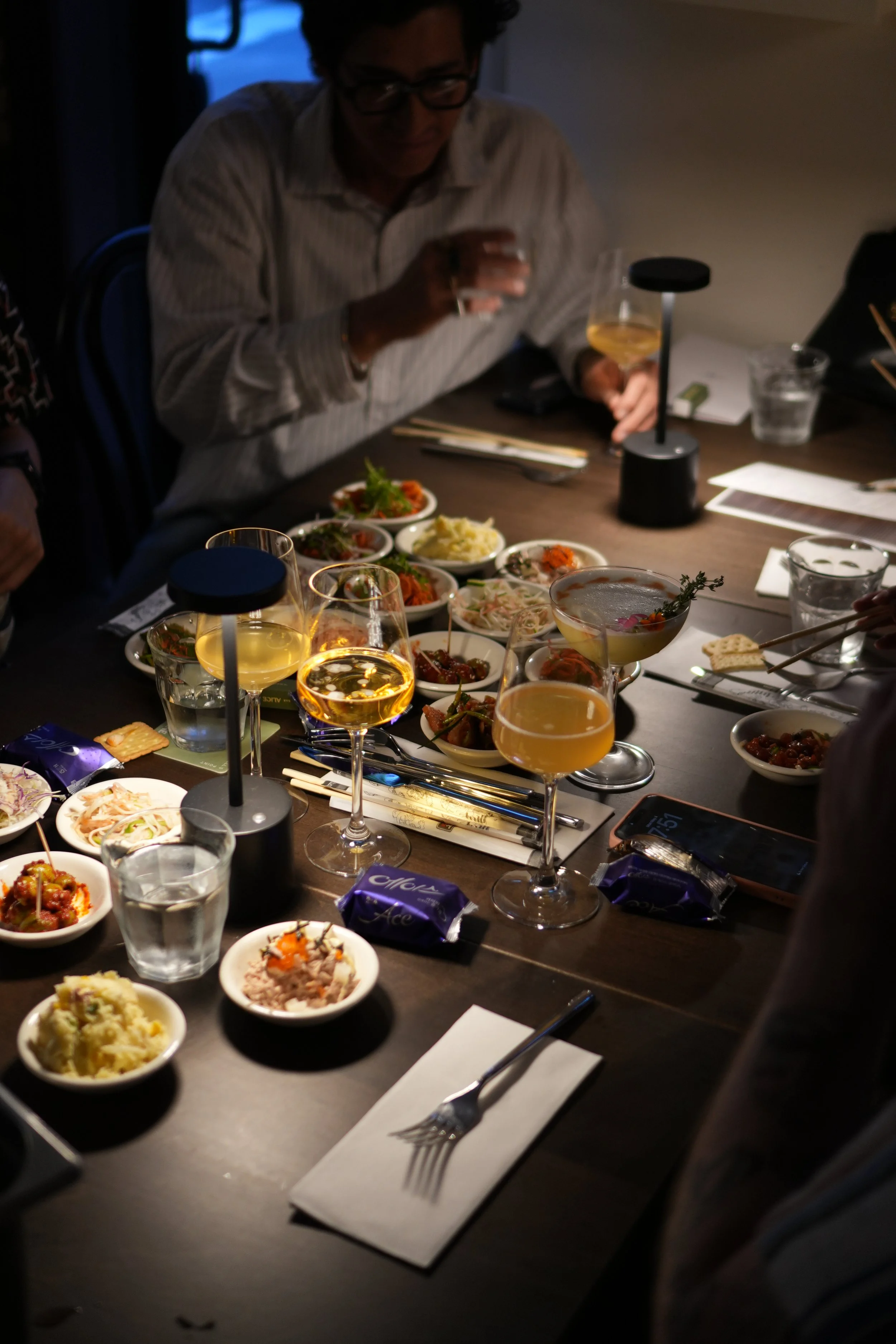 A dinner scene featuring a variety of Asian dishes in small bowls on a dark table, with glasses of white wine, a lit table lamp, and a person in the background holding a drink in Bar Alice Anne