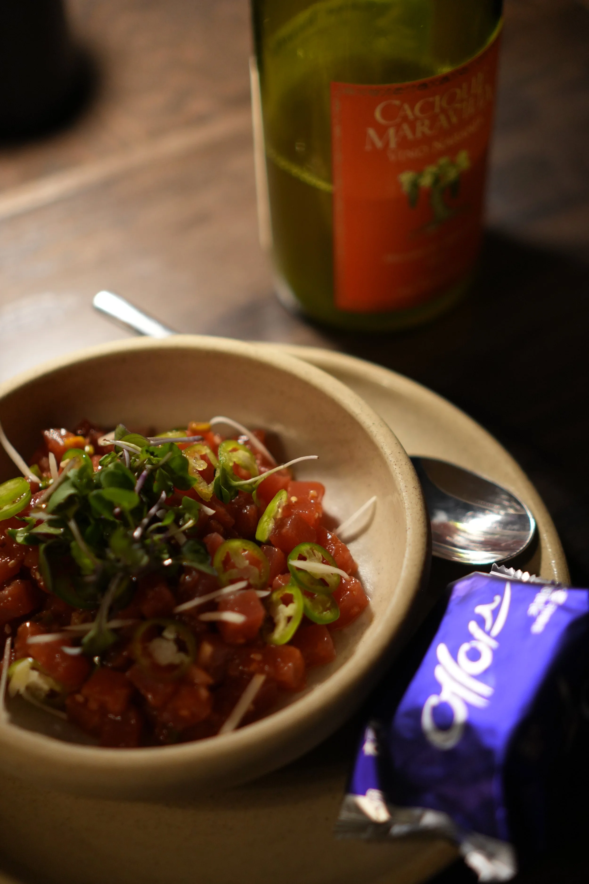 A bowl of chopped tuna tartare topped with microgreens, a fork, a bag of Ace crackers, and a bottle of orange natural wine in a bar.