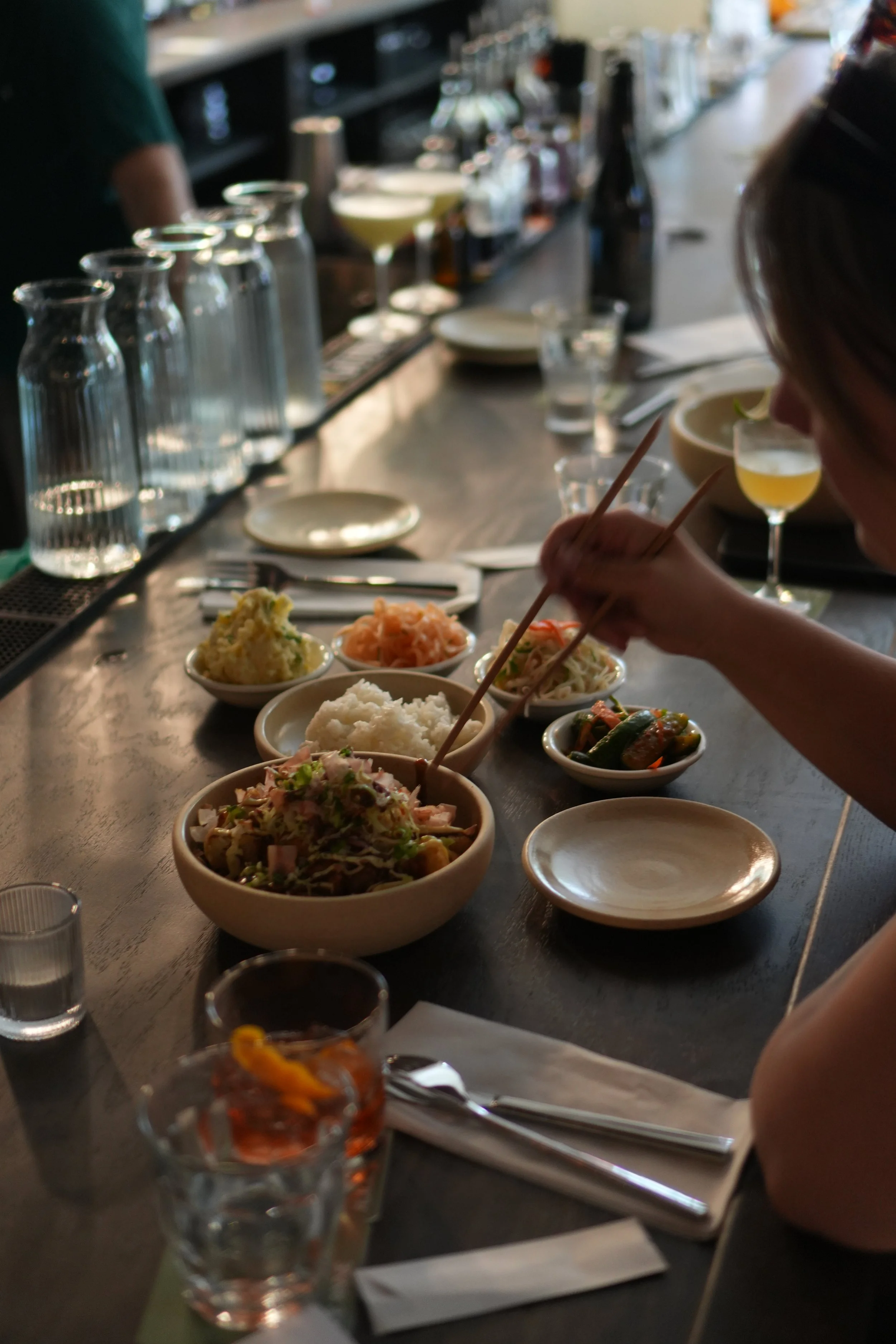 A person using chopsticks to eat a bowl of salad at a dining table with several small bowls of various dishes, glasses of water, and a bar counter with bottles in the background.