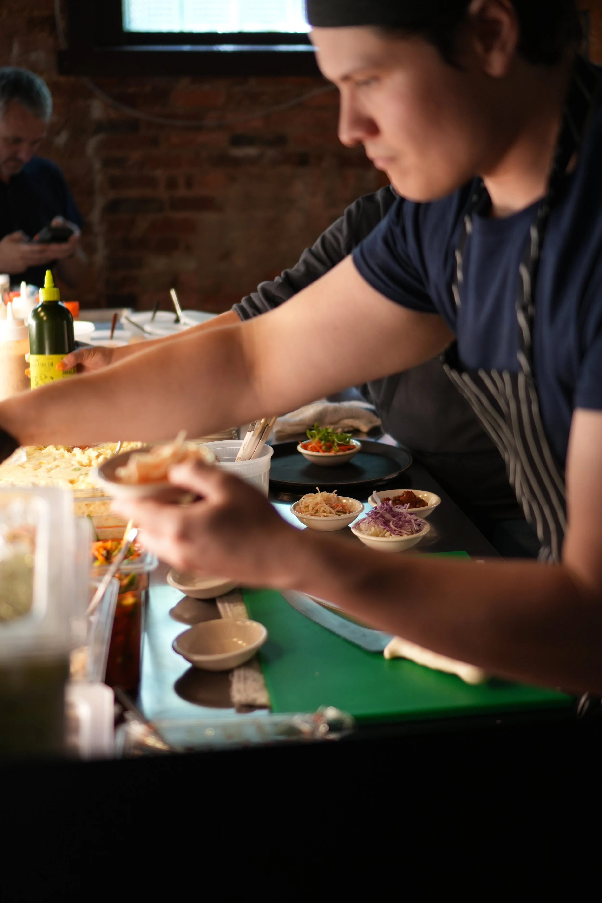 A person preparing Korean food at a counter with various small bowls of banchan, toppings and ingredients. In the background is a brick wall and a window above.