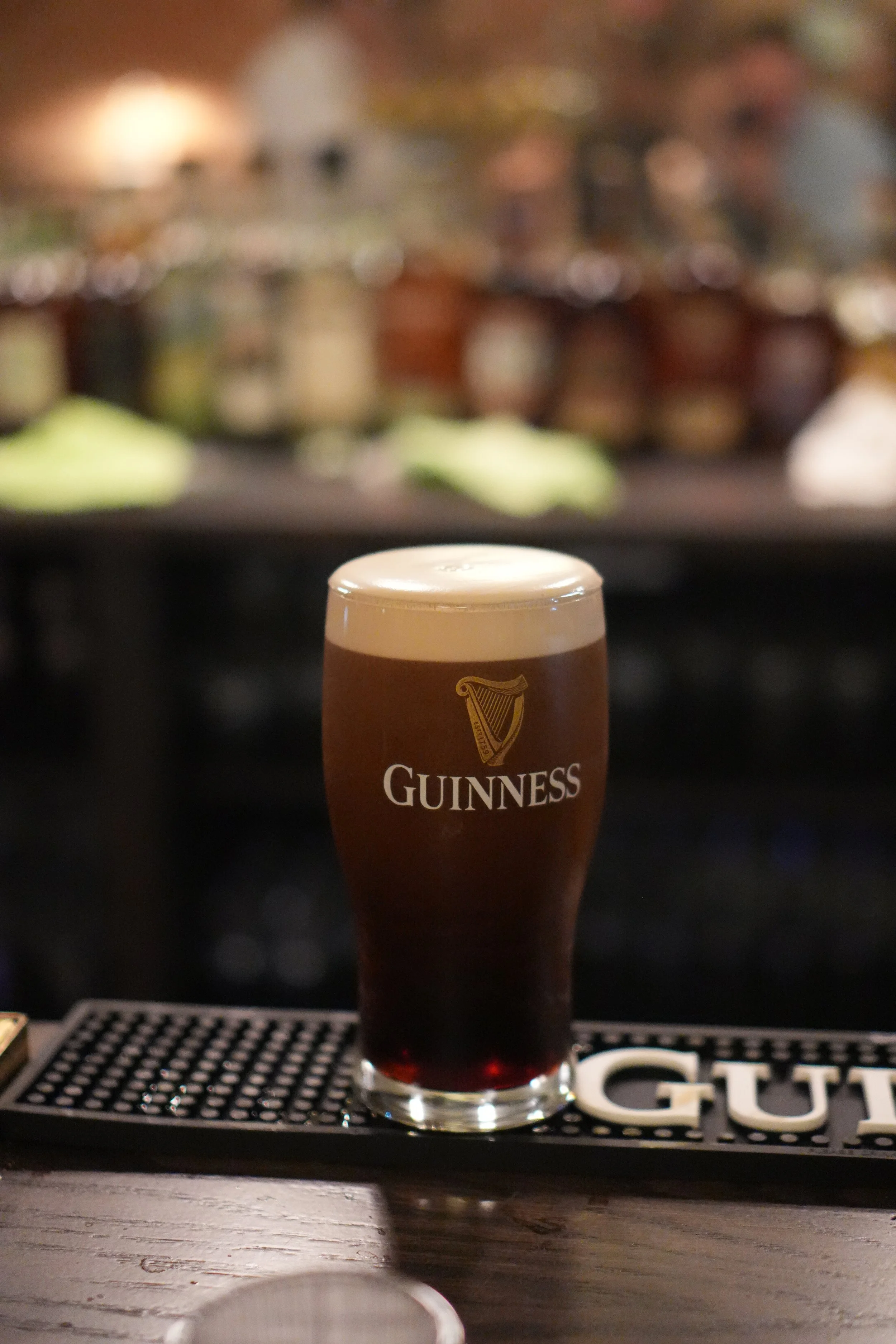 A pint of Guinness beer on a bar counter with a blurred background of people at Bar Alice Anne.
