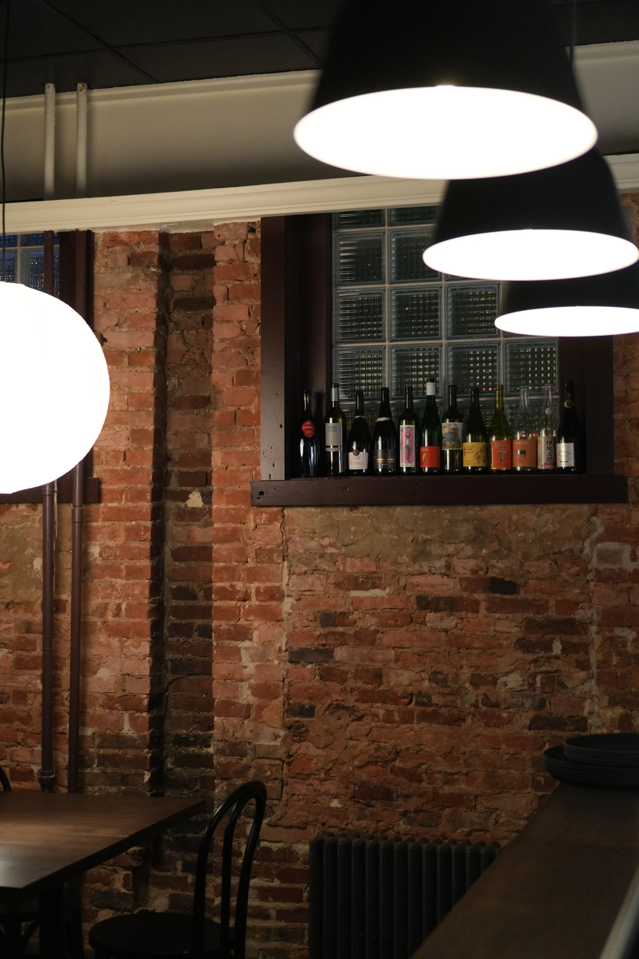 View of Bar Alice Anne with exposed brick walls, hanging pendant lights, and a shelf with wine bottles. Part of a table and chairs are visible in the foreground.
