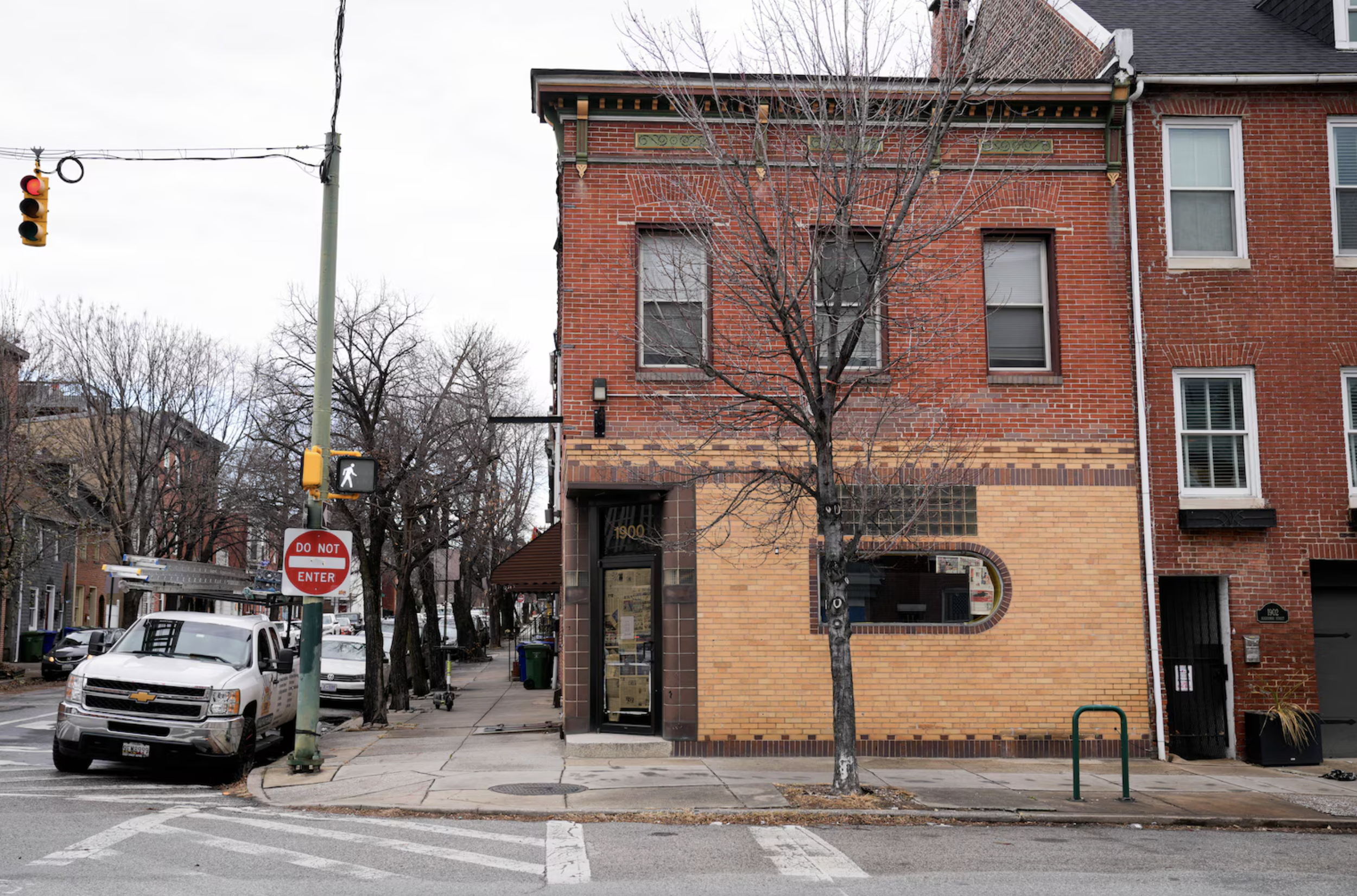 City street corner with a brick building on the right, leafless trees lined along the sidewalk, a white Chevrolet truck parked at the curb, traffic light with red and green signals overhead, and a do not enter sign on the pole. Overcast sky.